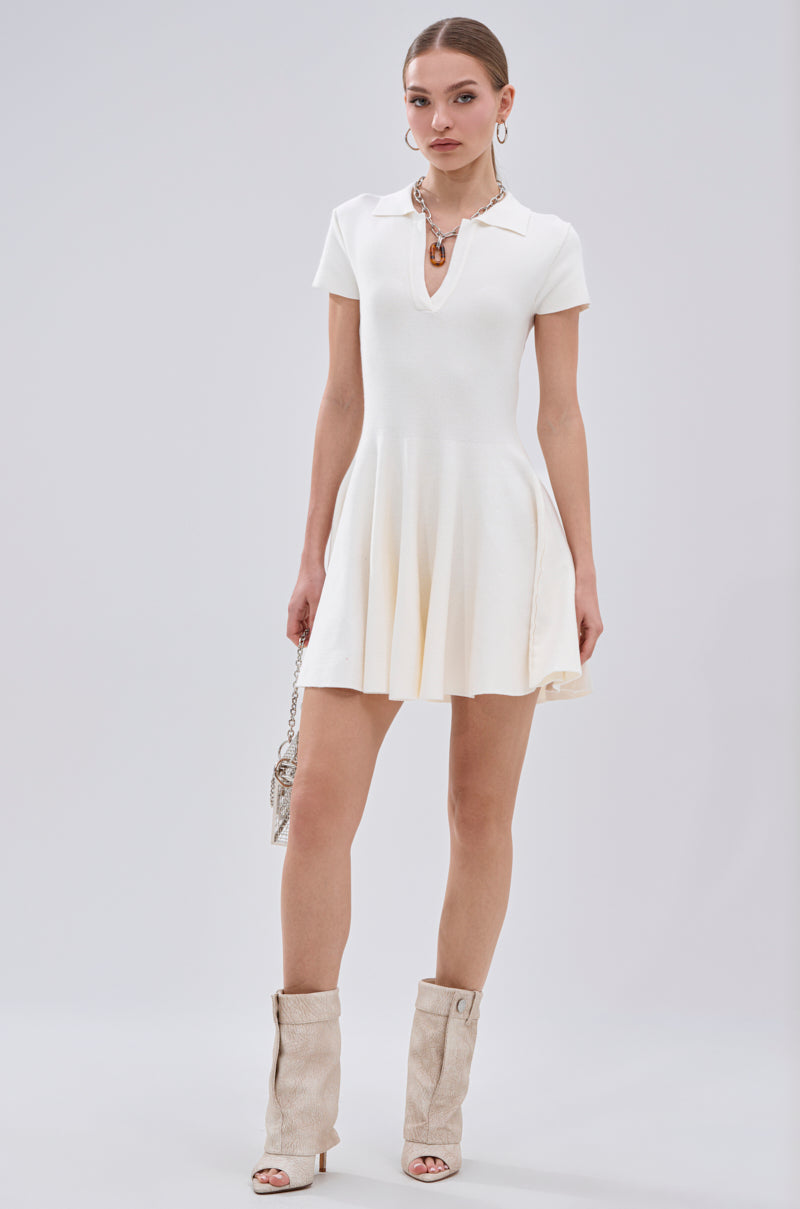 A woman poses against a plain backdrop in the SLEEK AND CHIC KNIT MINI DRESS IN WHITE, styled with a chunky necklace, hoop earrings, a small shoulder bag, and beige slouchy heeled boots. 6