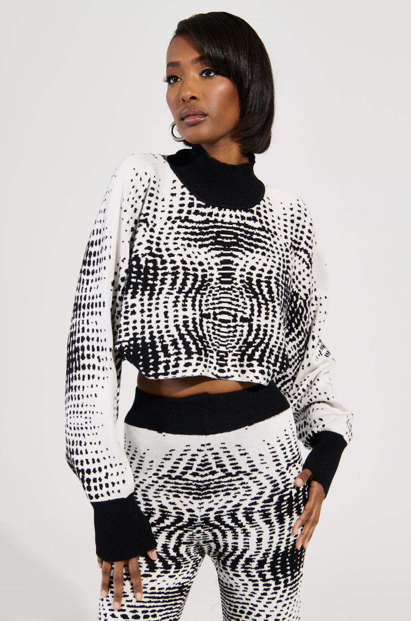 A woman stands confidently in the KALEIDOSCOPE DREAM PULL OVER SWEATER and matching pants, featuring an abstract wavy pattern. Her outfit's cropped silhouette complements her short hair as she poses with one hand on her hip against a plain background.