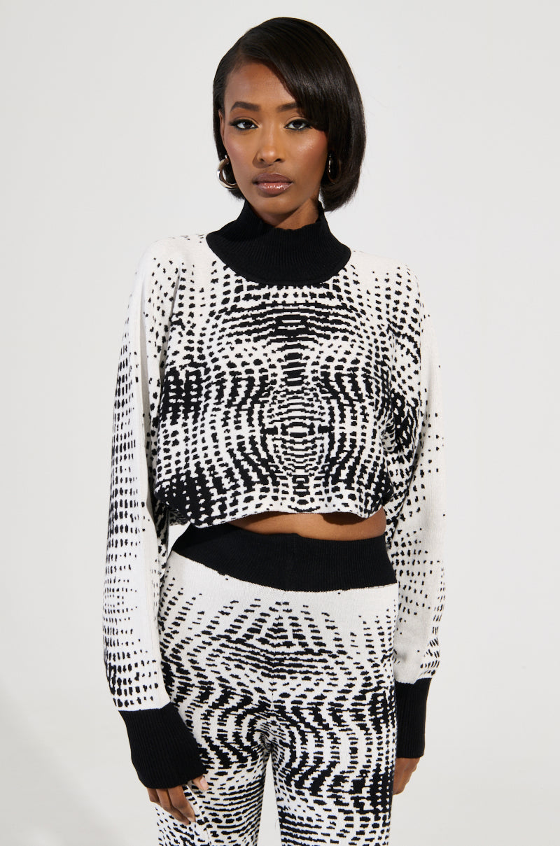 Wearing the KALEIDOSCOPE DREAM PULL OVER SWEATER and matching pants, an individual showcases a bold abstract wavy design. The cropped silhouette of the turtleneck sweater lends an edgy touch against a simple light backdrop, creating an effortlessly stylish impression.