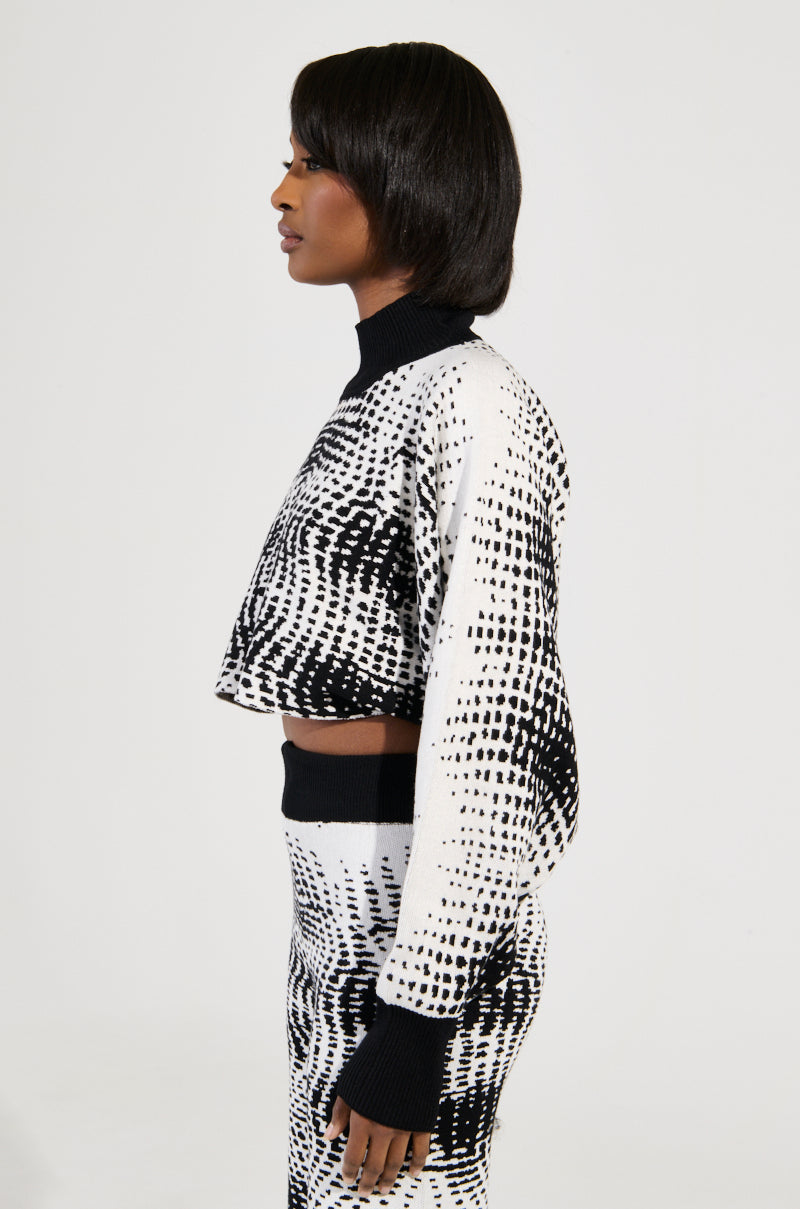 A person with short dark hair is wearing a matching black and white KALEIDOSCOPE DREAM PULL OVER SWEATER and skirt. They are standing in profile against a plain white background, their ensemble boasting an abstract wavy pattern that adds depth to the otherwise minimal setting.