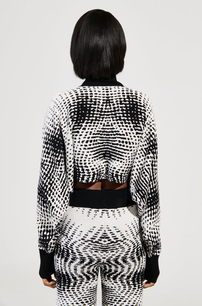 A person with short black hair wears a KALEIDOSCOPE DREAM PULL OVER SWEATER paired with matching pants, showcasing an abstract wavy pattern. The unique cutout design on the back adds interest to the ensemble. The plain, light-colored background highlights the outfit's bold statement.