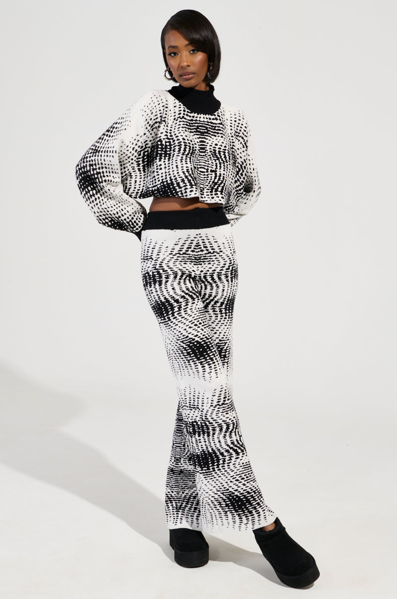 A person stands against a plain background, showcasing a KALEIDOSCOPE DREAM PULL OVER SWEATER with an abstract black and white wavy pattern. The outfit is paired with a fitted skirt and black platform shoes, while their hair is styled to one side, complementing the design's rhythm.