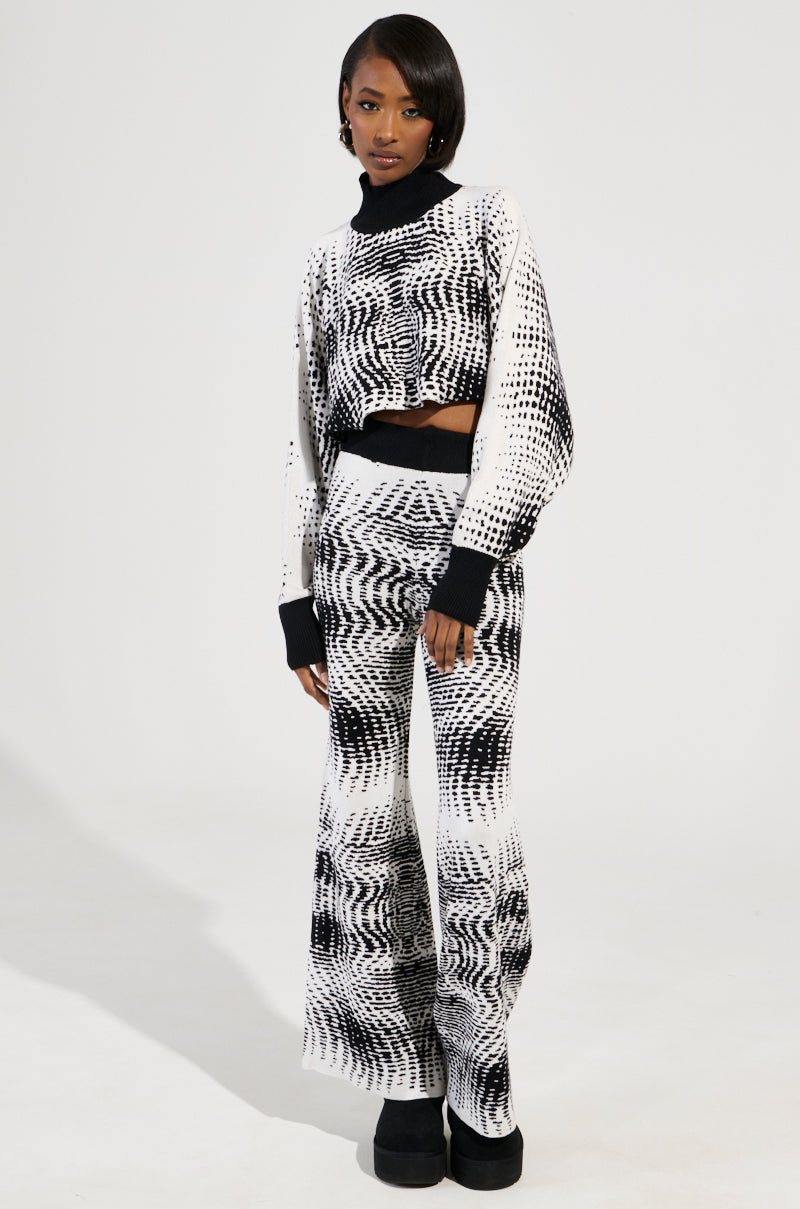 A person stands wearing the KALEIDOSCOPE DREAM PULL-OVER SWEATER, featuring a black and white abstract wave design akin to a kaleidoscope dream, paired with flared pants and black platform shoes against a plain white background.