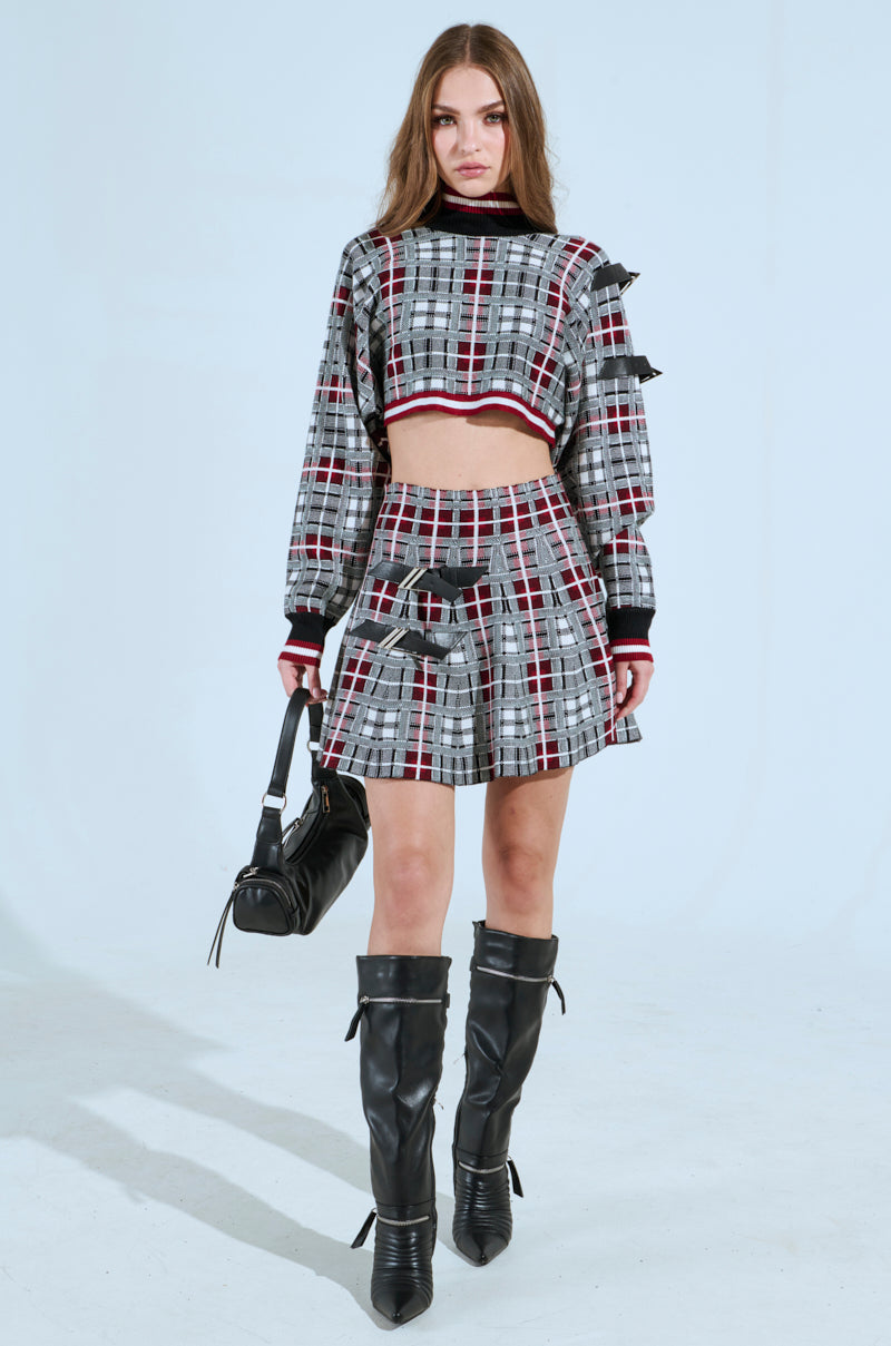 Amid a light blue backdrop, a person models punk fashion in the ALL EYES ON ME Buckle Detail Knit Mini Skirt in red paired with a matching cropped top. Knee-high black boots and a black handbag complete this rebellious look.
