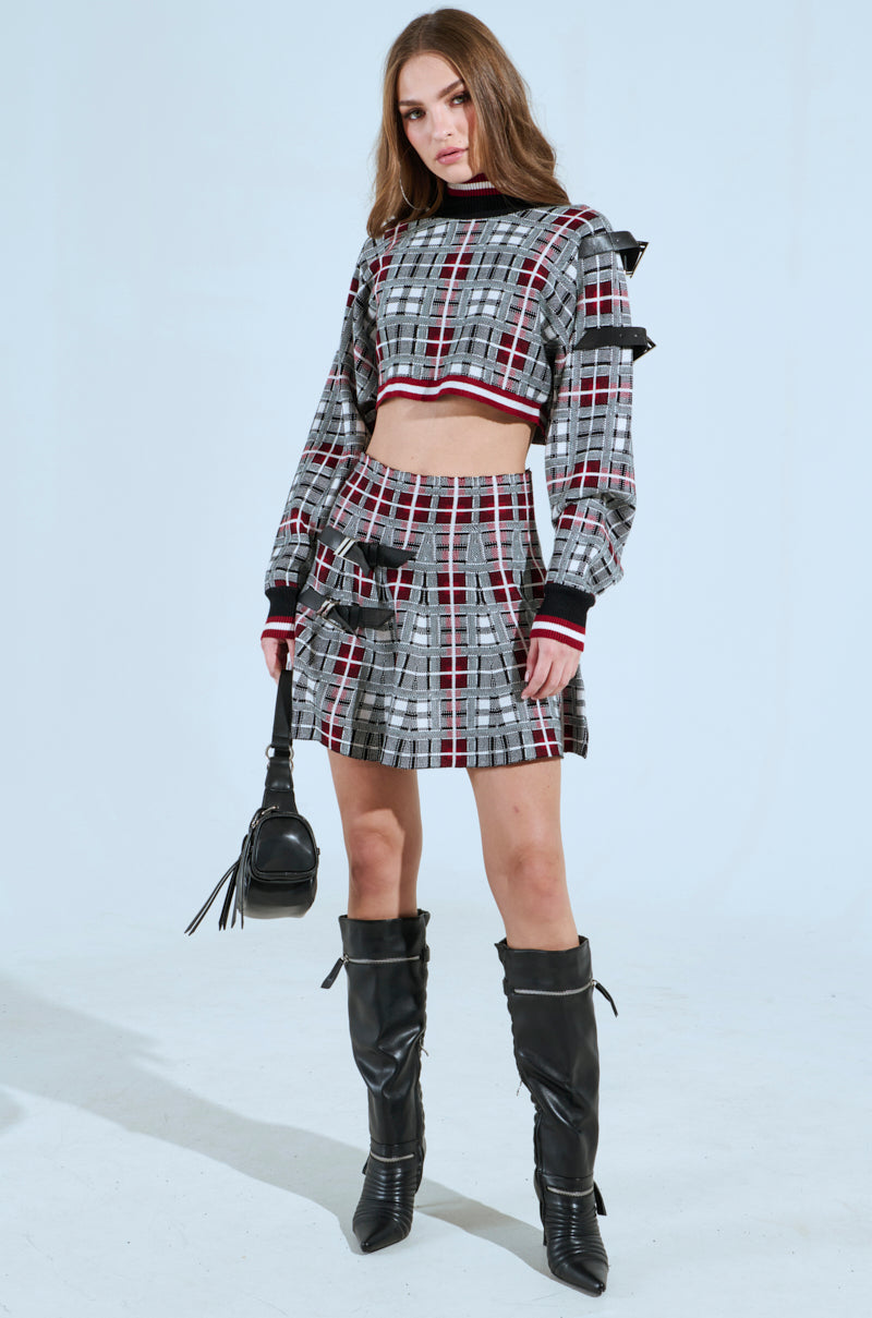 A person with long hair stands confidently in a punk fashion ensemble, featuring the "ALL EYES ON ME BUCKLE DETAIL KNIT MINI SKIRT IN RED" with ruffle details paired with a plaid long-sleeve crop top. They carry a black handbag and wear knee-high black boots against a minimalist, light-colored backdrop.