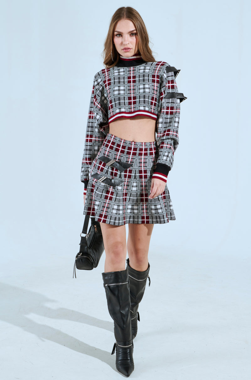 A woman exudes punk confidence in a bold ensemble featuring the "All Eyes on Me Buckle Detail Knit Mini Skirt in Red," paired with a cropped top, black knee-high boots, and a small black handbag against a light blue backdrop.
