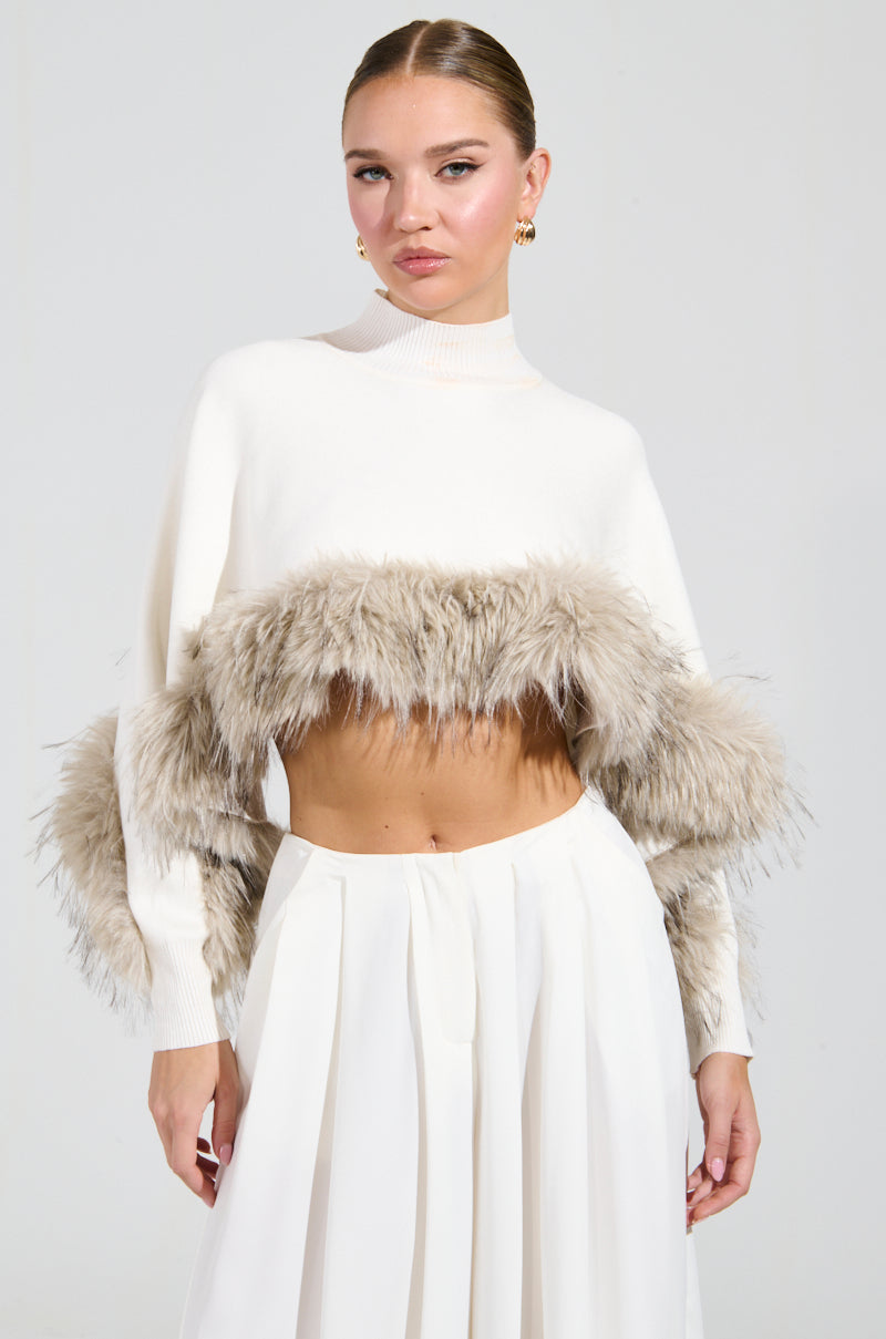 A person in the ABOUT DAMN TIME SWEATER, featuring a cropped fit with faux fur trim, pairs it stylishly with a pleated skirt against a simple backdrop. Their hair is pulled back and they accessorize with gold hoop earrings, adding a hint of glamour to the ensemble.