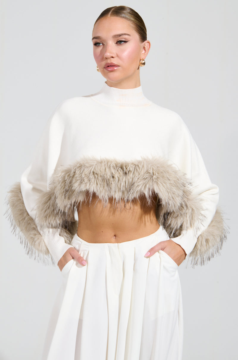A person poses confidently in the ABOUT DAMN TIME SWEATER, a chic white cropped sweater with fur trim, paired with a pleated skirt. Gold earrings add a touch of elegance to their look as they rest their hands casually in their pockets against the plain gray background.