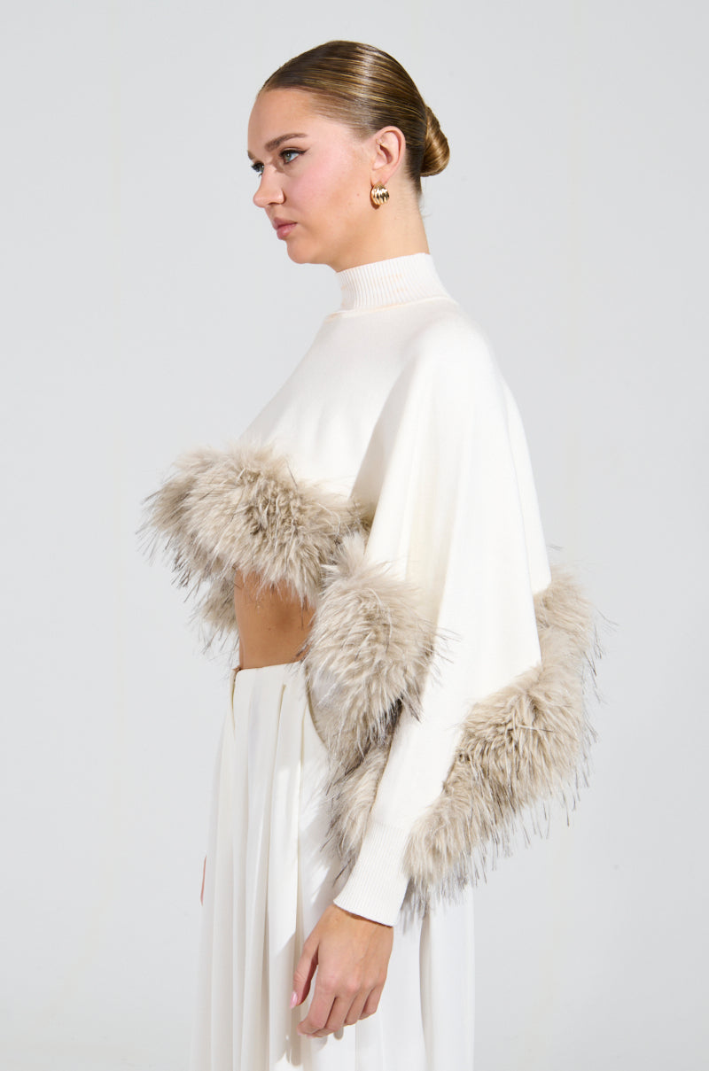 A person with their hair in a sleek bun wears a white outfit featuring feathered accents on the top, paired with the ABOUT DAMN TIME SWEATER in a cropped fit. They have gold hoop earrings and are looking to the side against a plain background.