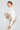 A person with their hair in a sleek bun wears a white outfit featuring feathered accents on the top, paired with the ABOUT DAMN TIME SWEATER in a cropped fit. They have gold hoop earrings and are looking to the side against a plain background.