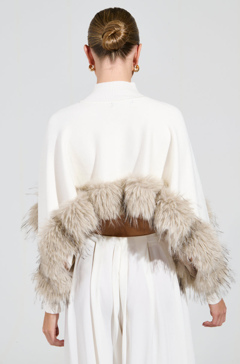 A person with their hair in a bun wears the ABOUT DAMN TIME SWEATER, which is a white turtleneck accented by a beige, furry trim on the back. They pair it with white pants and large hoop earrings while standing against a plain background.