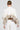 A person with their hair in a bun wears the ABOUT DAMN TIME SWEATER, which is a white turtleneck accented by a beige, furry trim on the back. They pair it with white pants and large hoop earrings while standing against a plain background.