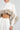 Wearing the ABOUT DAMN TIME SWEATER, which features a chic white turtleneck crop top design with fluffy, light brown fur accents, a person achieves a stylish cropped fit. They complete the look with a high-waisted, pleated white skirt against a plain and minimalistic background.
