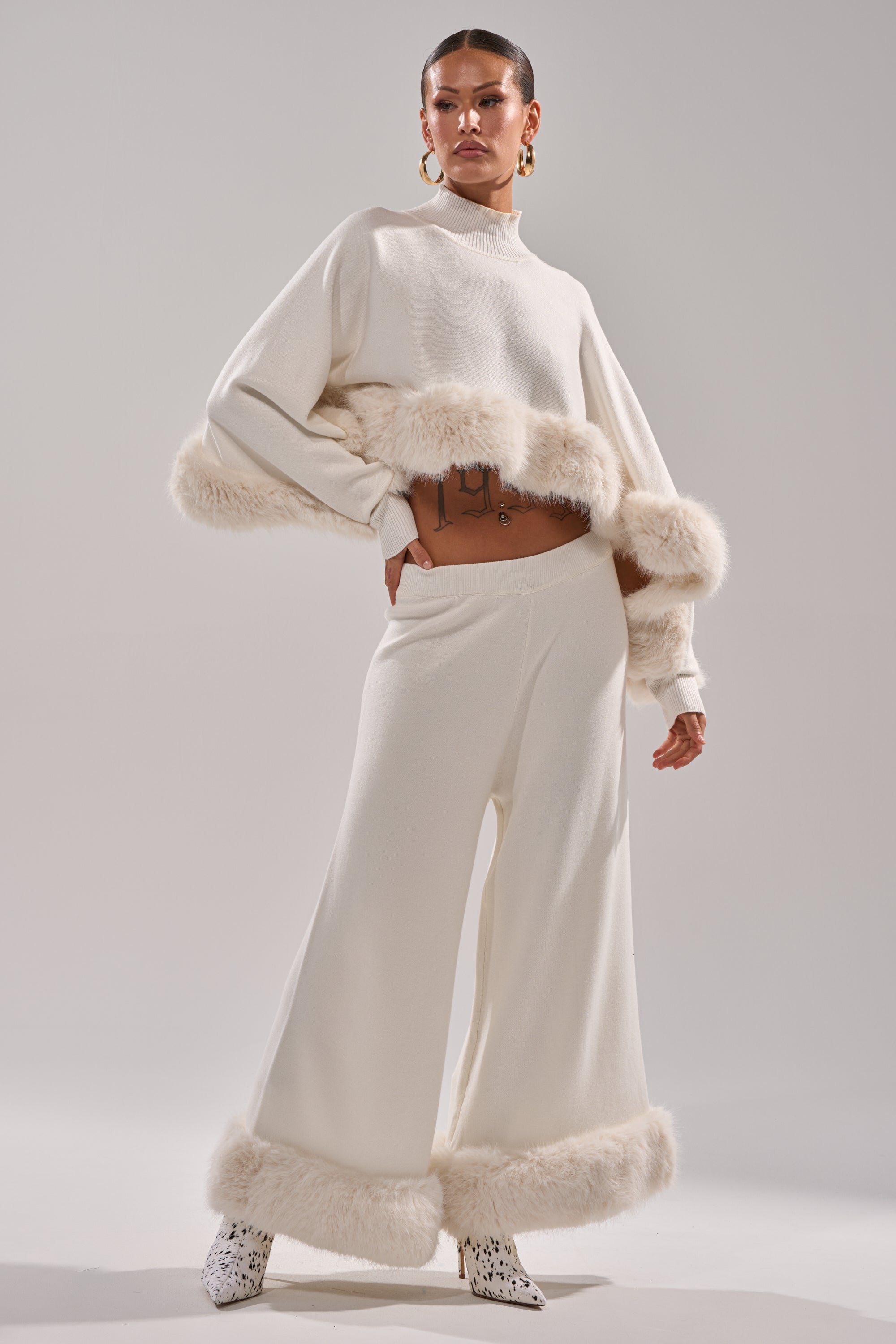 A woman models the QUIET LUX FUR TRIM SWEATER PANTS IN WHITE—cream knit wide-leg pants and long-sleeve top with white fur trim, styled with patterned ankle boots and gold hoop earrings against a simple background. 0