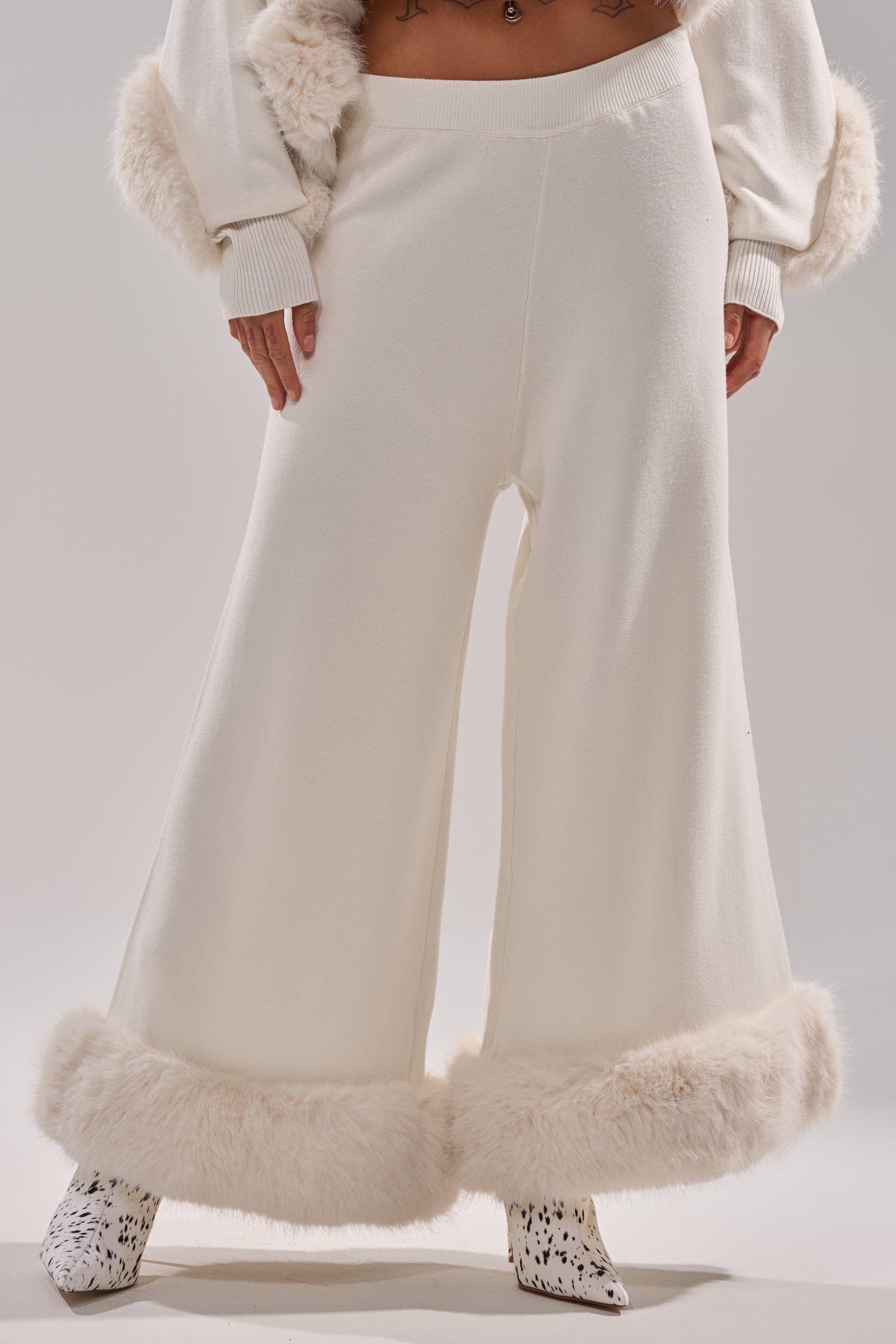 Wearing the QUIET LUX FUR TRIM SWEATER PANTS IN WHITE—wide-leg pants with faux fur ankle trim—styled with a matching sweater and white pointed boots with black speckles. Only the lower body is visible. 1
