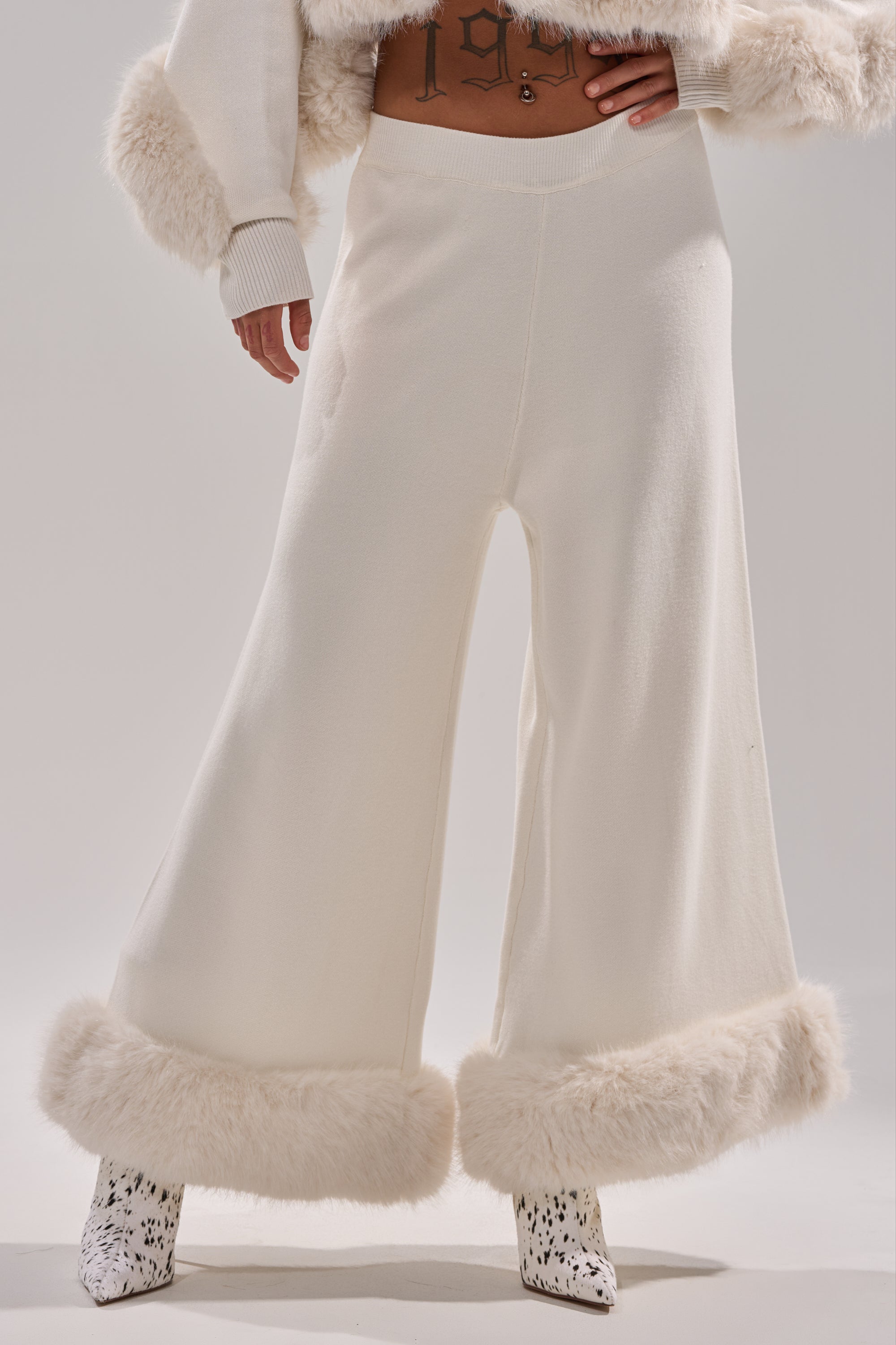Model in QUIET LUX FUR TRIM SWEATER PANTS IN WHITE—wide-leg white pants with faux fur trim at the hem—paired with a matching fur-trimmed sweater and spotted boots. Midriff and tattoo visible; light-colored background. 2