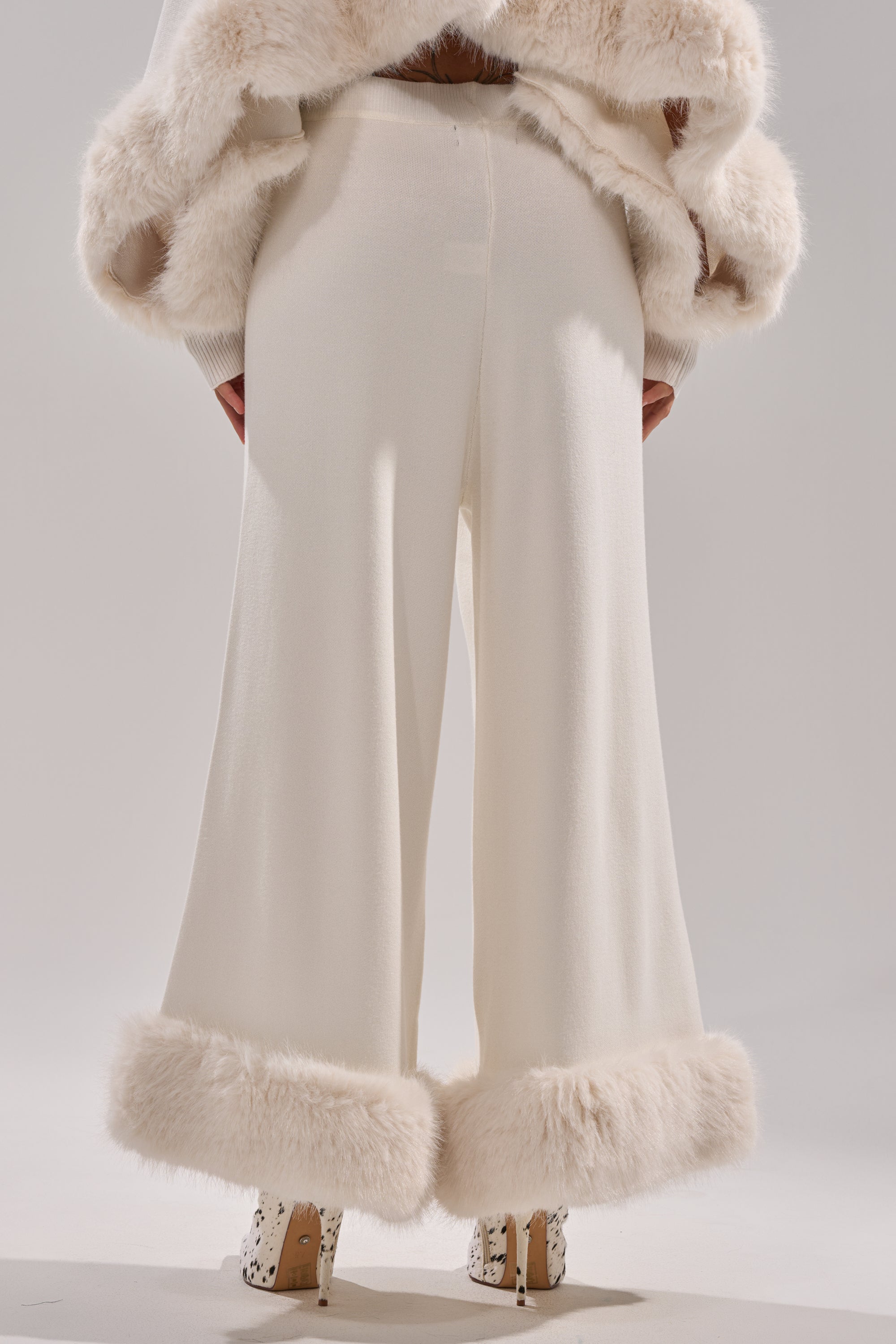 A person faces away, wearing QUIET LUX FUR TRIM SWEATER PANTS IN WHITE paired with a matching fur jacket and white shoes with a black spotted pattern, set against a plain light background. 4