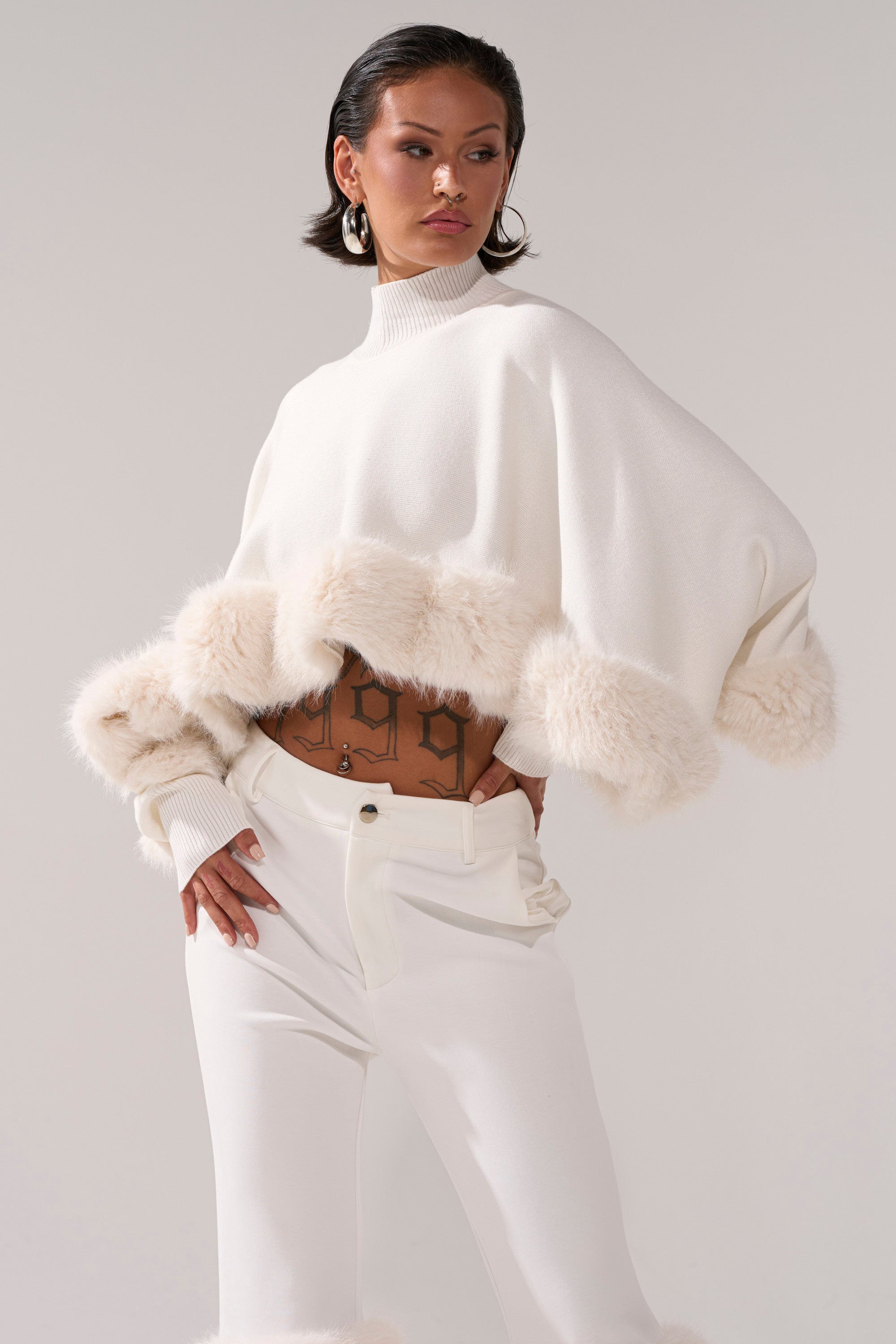 A person confidently poses in the QUIET LUX FUR TRIM SWEATER IN WHITE, a cream cropped poncho with fur trim, paired with high-waisted pants that reveal tattoo designs on their midriff.