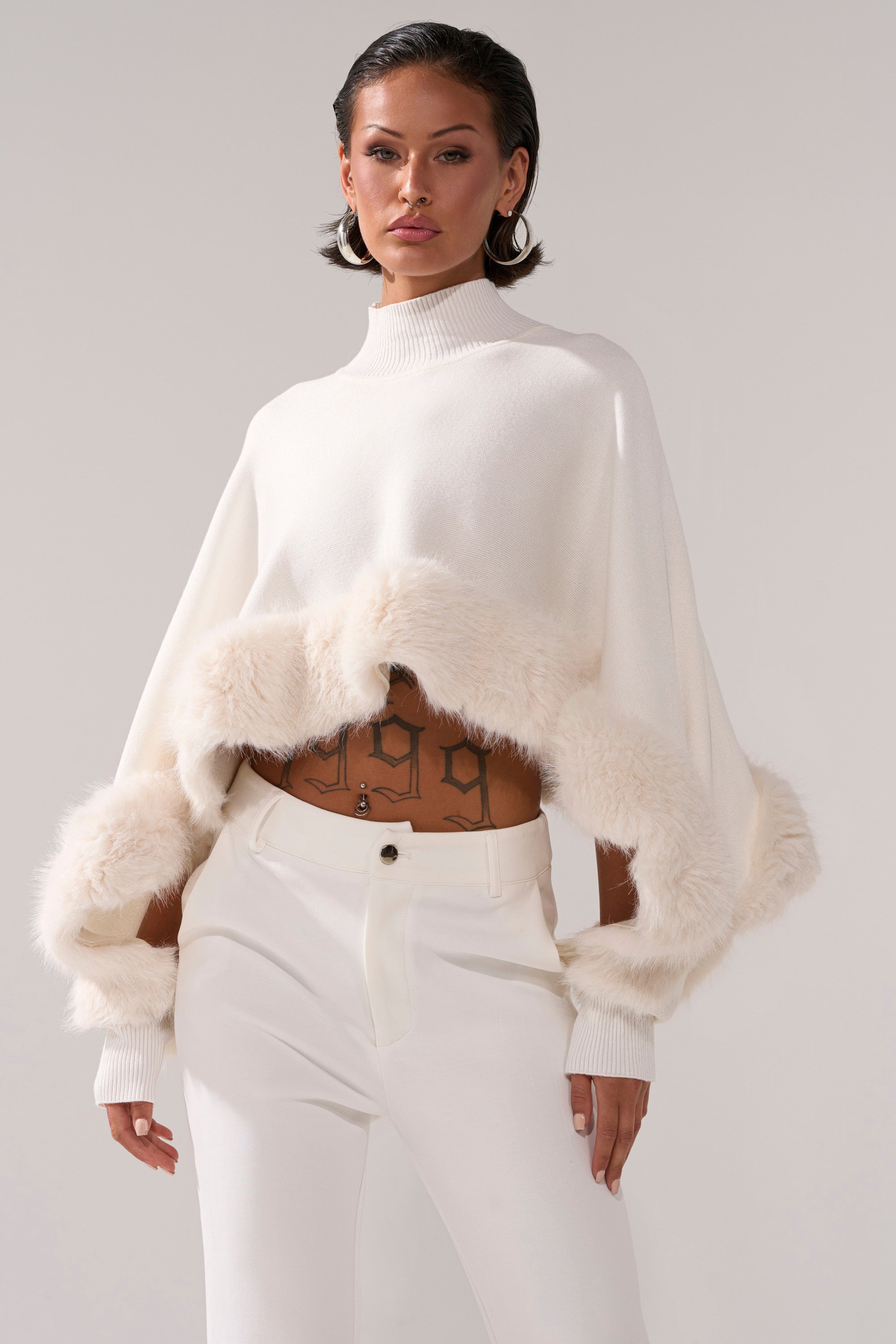 A woman wears the QUIET LUX FUR TRIM SWEATER IN WHITE with matching pants, showing off midriff tattoos and short, styled hair against a neutral background.