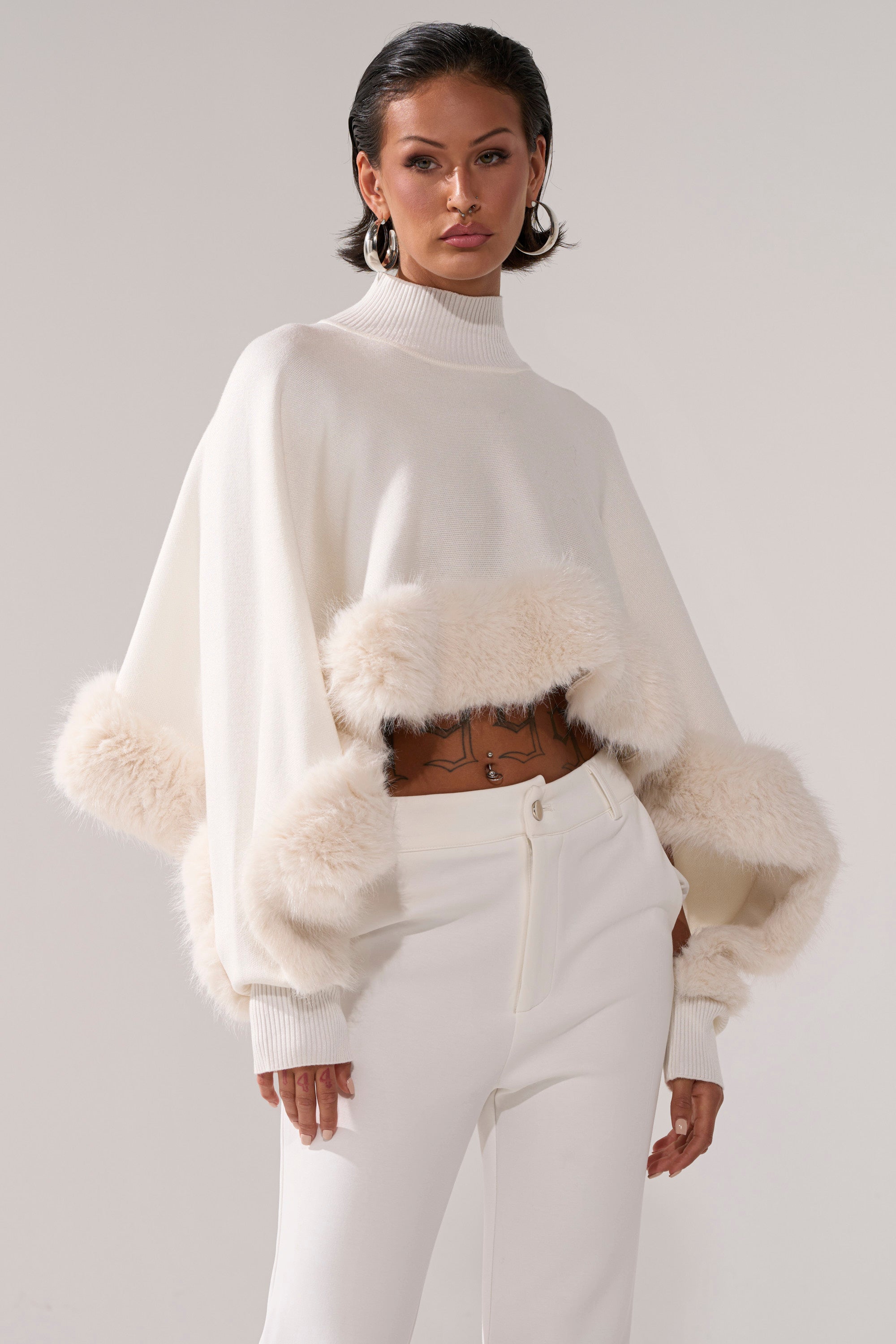 A woman models the QUIET LUX FUR TRIM SWEATER IN WHITE, a minimalist knit with fluffy waist and sleeve trim, paired with high-waisted white pants against a neutral background.