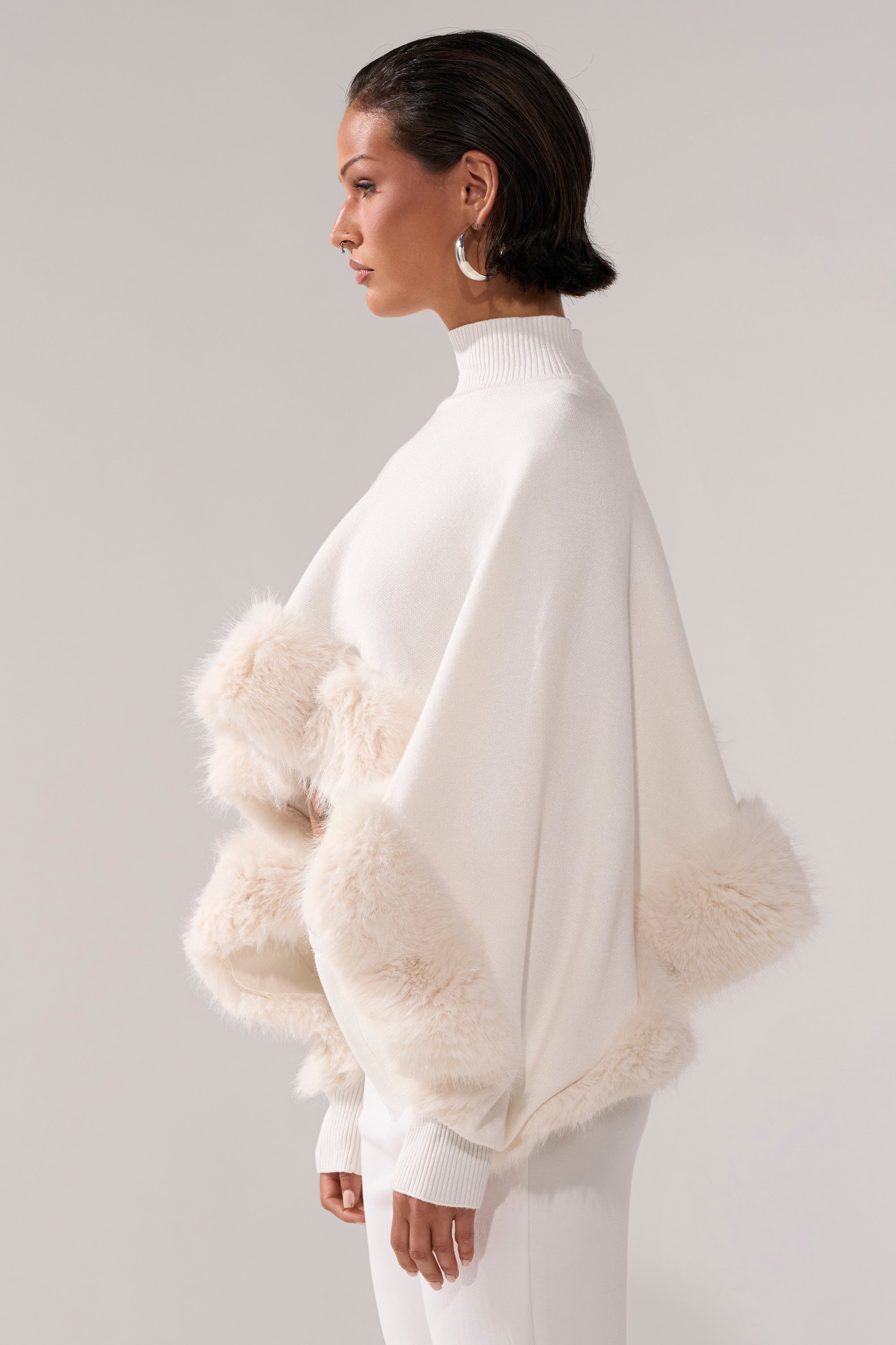 A woman in profile models the QUIET LUX FUR TRIM SWEATER IN WHITE, styled as a cape with white pants and gold hoop earrings, her short hair sleek against a plain light background.