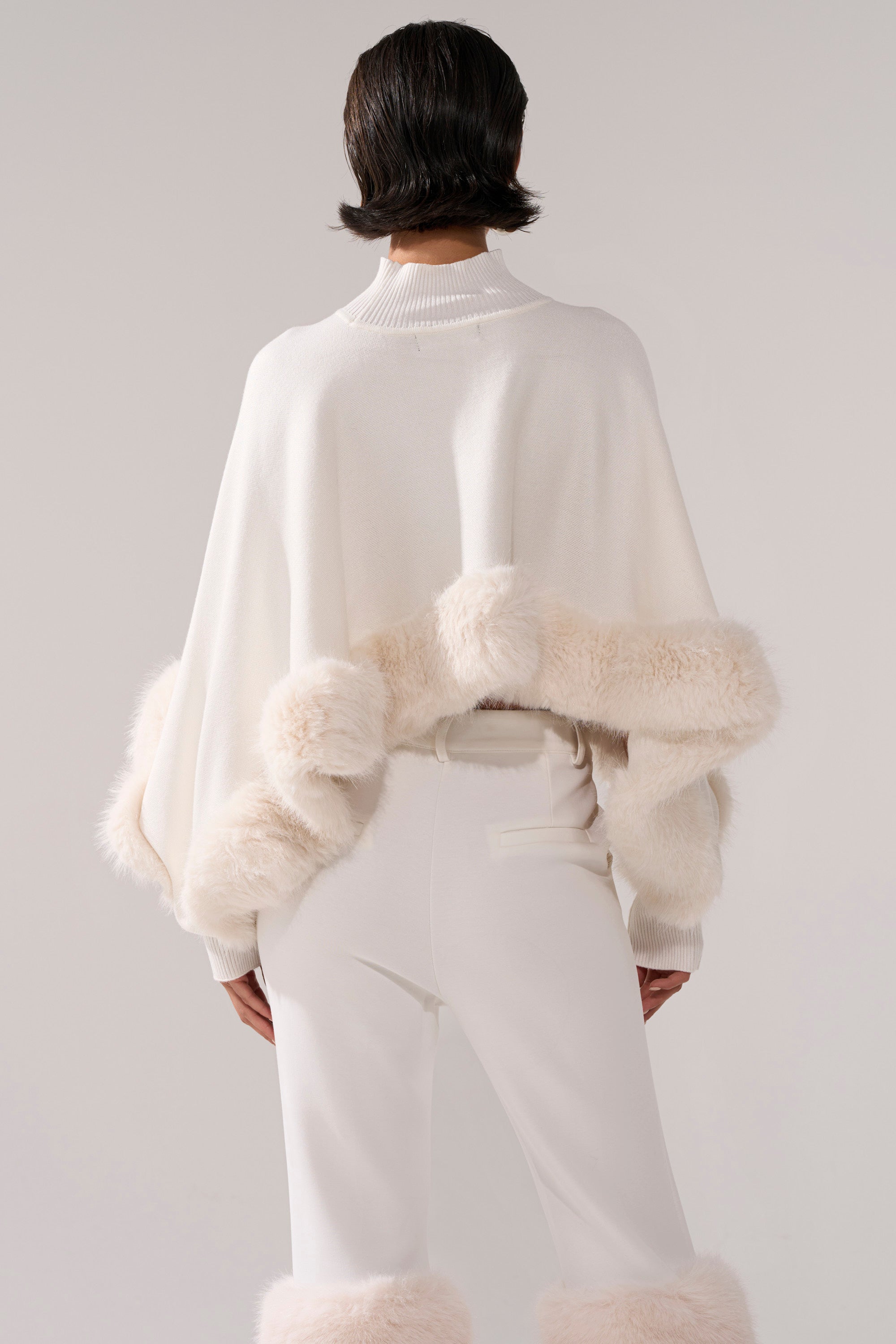 A person faces away, wearing the QUIET LUX FUR TRIM SWEATER IN WHITE styled as a minimalist cape with light beige fur trim, paired with matching white pants featuring fur accents at the ankles. The background is plain and light-colored.