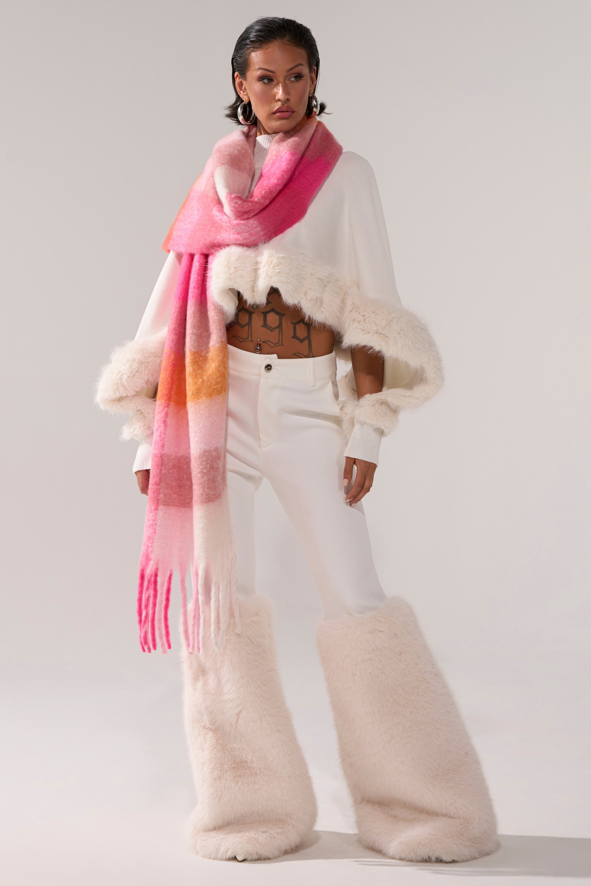 A woman poses confidently against a plain background wearing the QUIET LUX FUR TRIM SWEATER IN WHITE, paired with wide white faux fur pants, a sheer brown top, long pink gradient scarf, and hoop earrings.