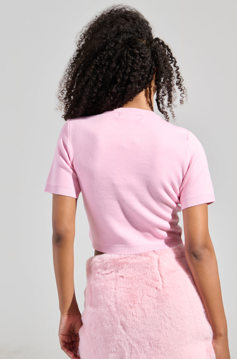 A person with curly hair stands facing away, showcasing a cropped silhouette that includes the "KEEP ME OUT THE LOOP TINY BOW SWEATER IN PINK," featuring short sleeves and paired with a pink fuzzy skirt. The stretchy knit outfit stands out against the plain, light-colored background.