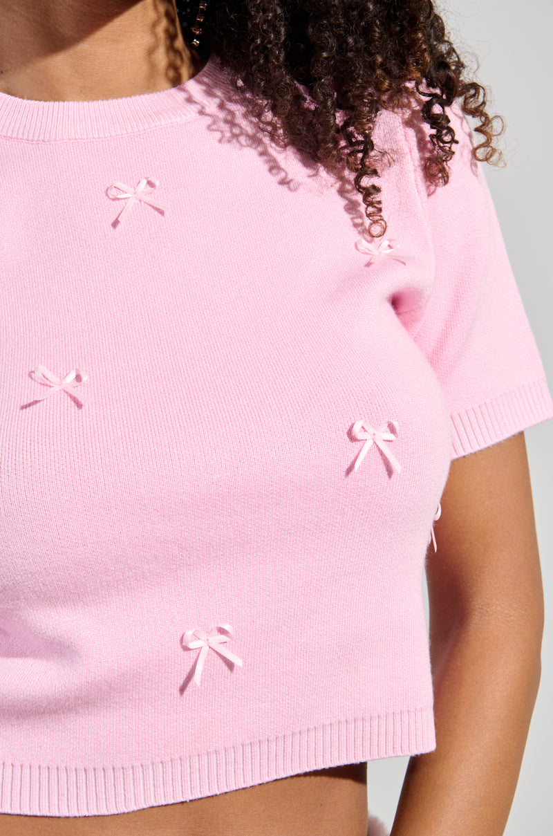 A person with curly hair wears the KEEP ME OUT THE LOOP TINY BOW SWEATER IN PINK, showcasing its cropped silhouette and small decorative bows. The image highlights the texture and details of the stretchy knit top against a neutral background.