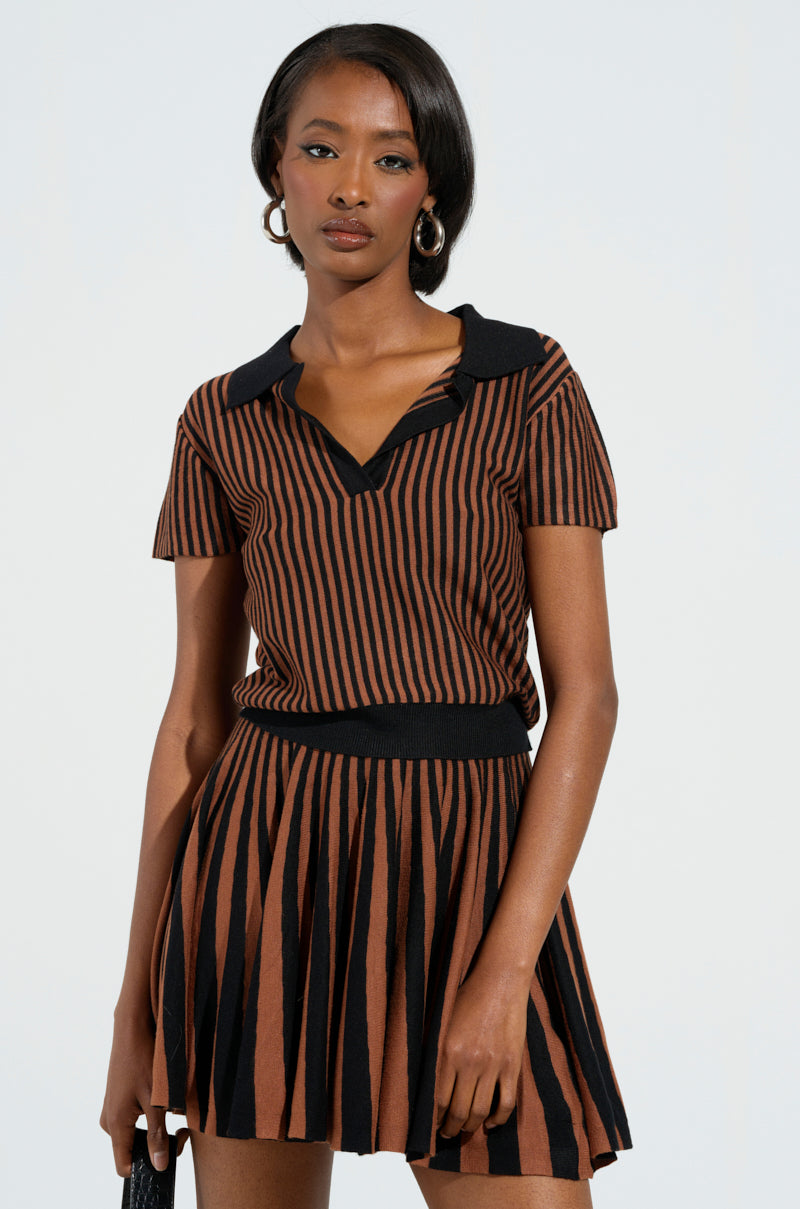 Wearing the GIRL HAS GAME POLO, a person stands against a plain background in a stretchy knit brown and black striped short-sleeved dress from the AKIRA Label, holding a small dark clutch. Their medium-length hair is styled neatly and paired with hoop earrings, exuding effortless elegance.