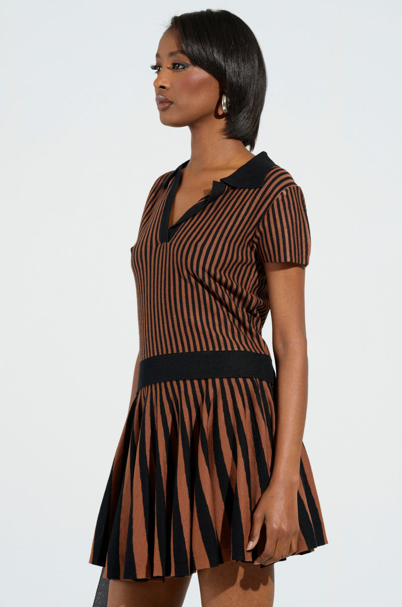 A woman wears the GIRL HAS GAME POLO from the AKIRA Label collection, a stylish short-sleeve dress with brown and black vertical stripes and a pleated skirt. The stretchy knit fabric enhances her medium-length dark hair as she looks to the side, highlighting the elegant deep V-neckline and fashionable collar.