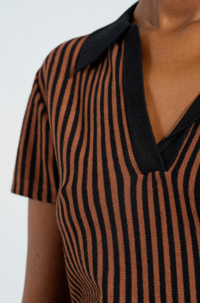 A person is wearing the GIRL HAS GAME POLO by AKIRA Label, showcasing vertical brown and black stripes with a contrasting black collar. The short-sleeved, V-neck polo is made from a stretchy knit fabric, all set against a light-colored backdrop.