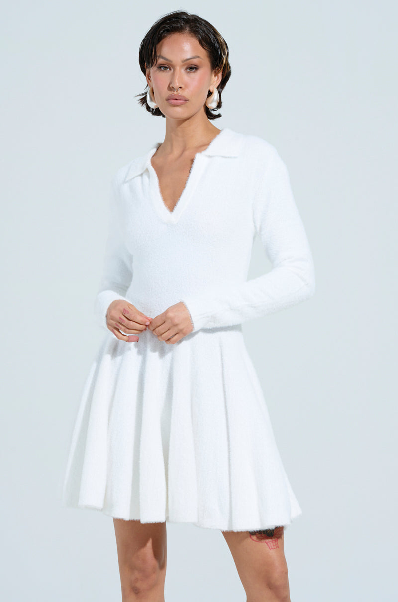 Against a light background, someone in the COZY GIRL CHIC FUZZY KNIT MINI DRESS WITH LONG SLEEVES stands, accessorized with statement earrings and short hair, epitomizing cozy girl chic.
