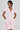A person in the BOW DOWN GIRL MINI DRESS IN PINK, featuring a flared skirt and small bow details, poses with hands behind their back. They have short dark hair and large pearl earrings, and the dress has a light pink color with a V-neck and collar.