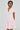 A woman wears the BOW DOWN GIRL MINI DRESS IN PINK, a light pink mini with a v-neck and bow details. The skater silhouette with a flared skirt complements her shoulder-length dark hair as she gazes to the side, set against a plain gray background.