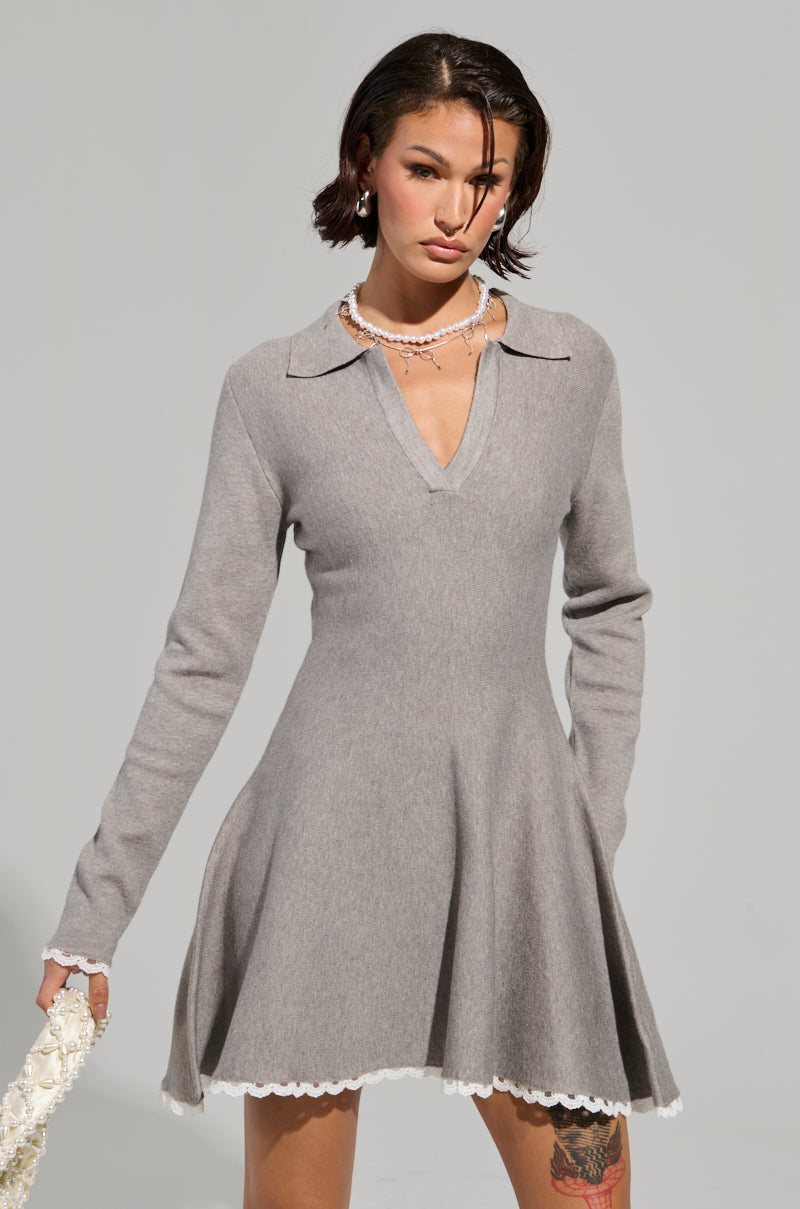 Dressed in the IN THE DETAILS KNIT MINI DRESS, which features a flared skirt and delicate white lace trim, a person holds a matching white, lacy accessory. The neutral gray background accentuates the dress's elegant simplicity.