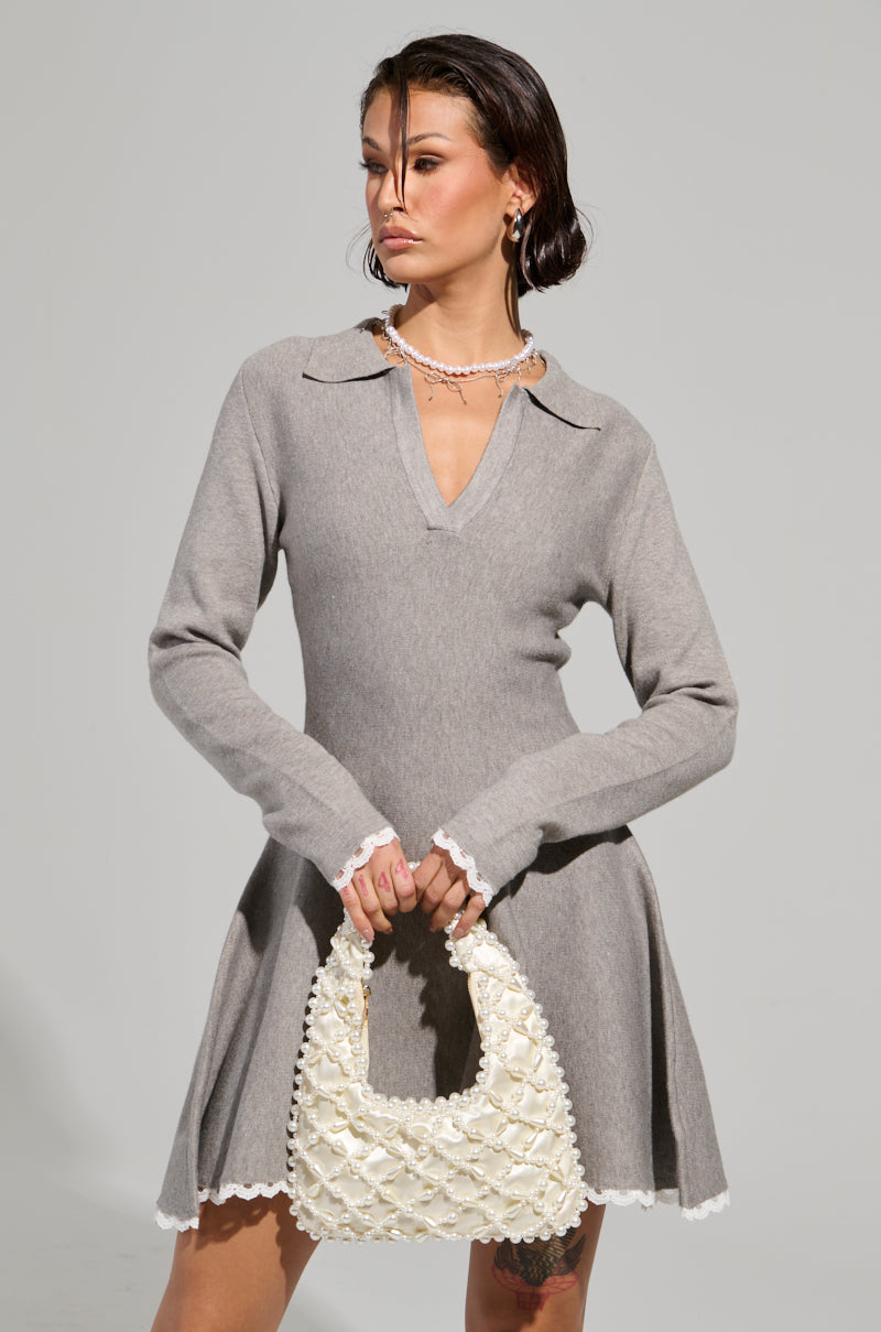 A person wearing the IN THE DETAILS KNIT MINI DRESS, which features a gray color, flared skirt, and V-neck collar, holds a textured cream handbag. They have short dark hair, wear a chain necklace, and stand against a plain background.
