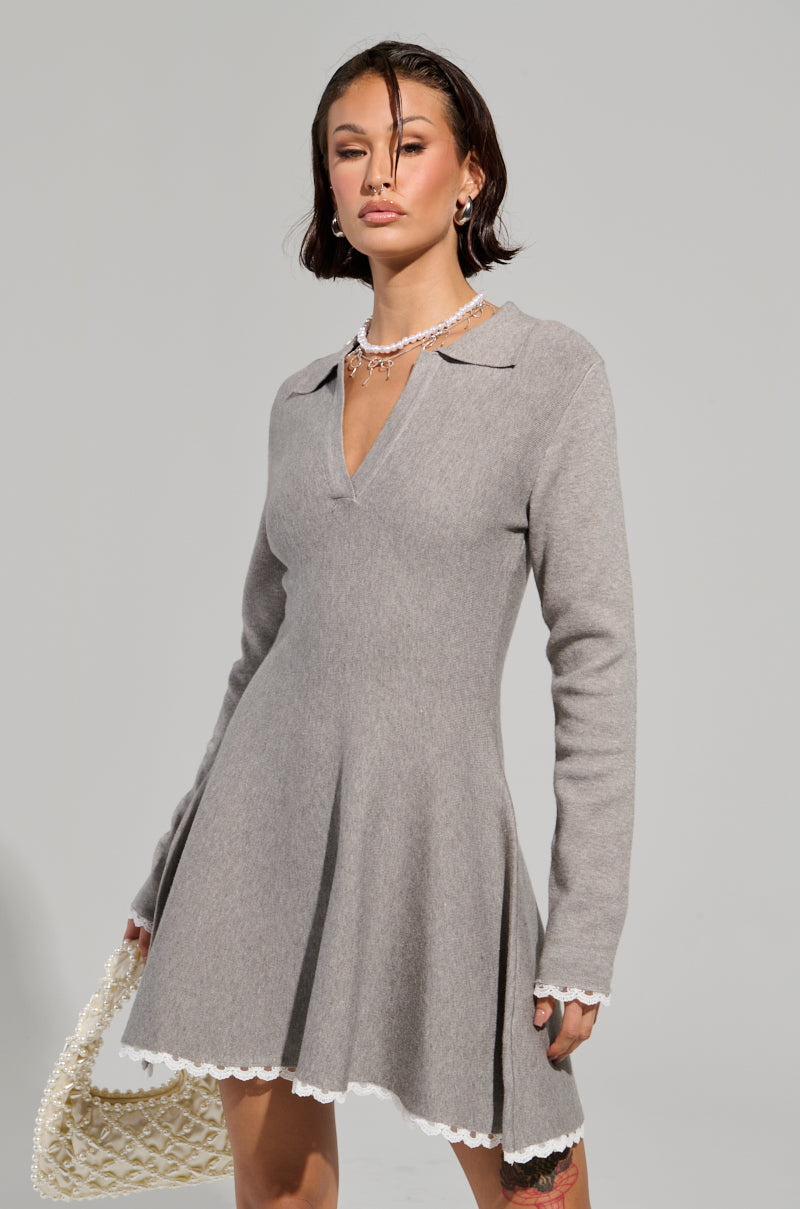 A person with short dark hair wears the IN THE DETAILS KNIT MINI DRESS, a long-sleeved gray fit 'n flare dress adorned with white lace trim. They hold a textured white handbag against the plain light gray background.