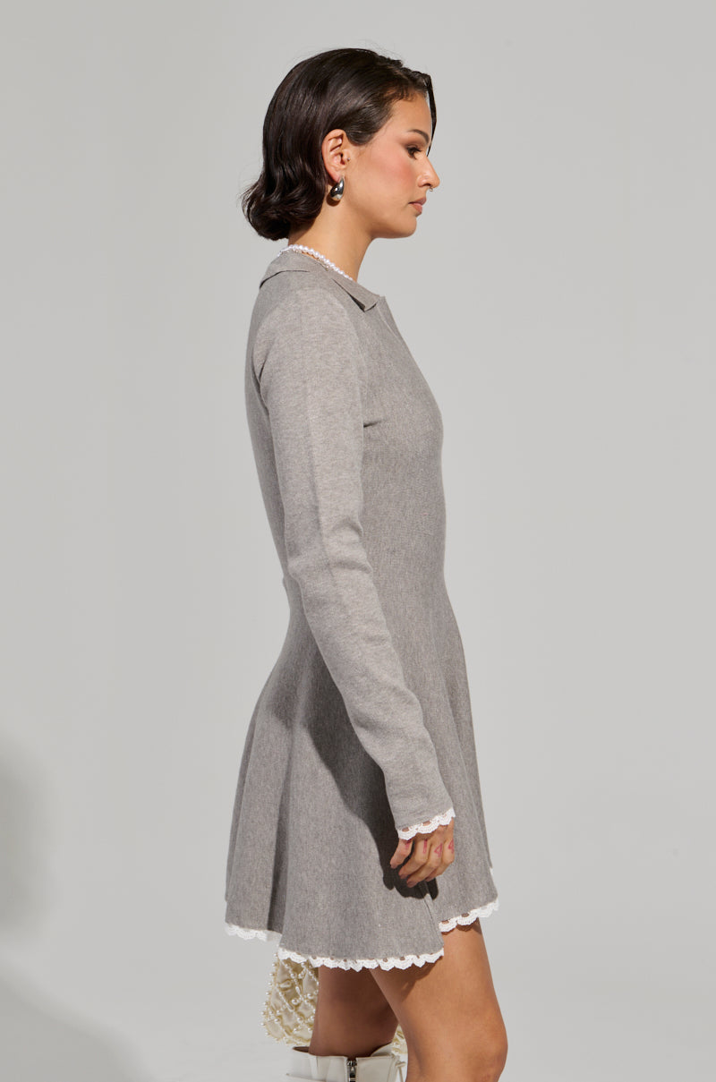 A person with short dark hair stands in profile, wearing the "In The Details" Knit Mini Dress. This gray dress features long sleeves and a fit 'n flare silhouette, beautifully highlighting its fitted waist with delicate lace trim against the muted gray background.