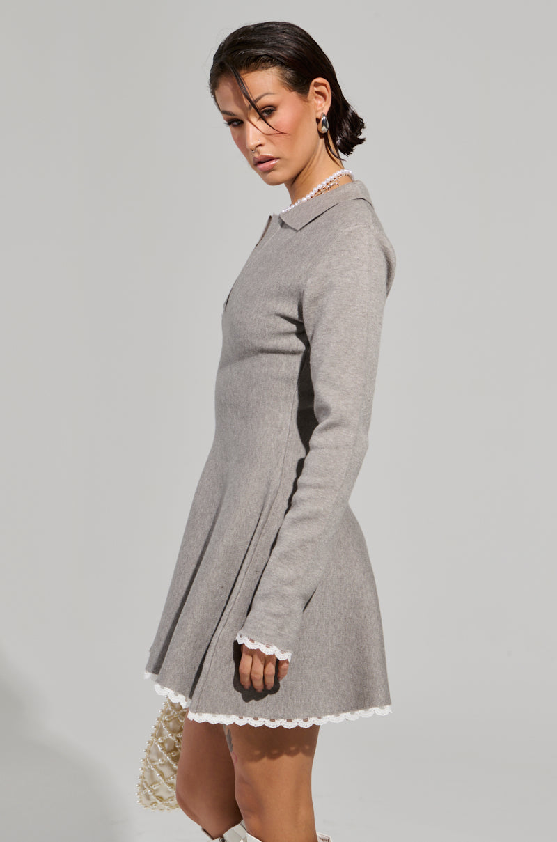 A person in an 'IN THE DETAILS KNIT MINI DRESS,' featuring a sophisticated gray fit 'n flare design with delicate lace trim, stands sideways, gazing at the camera. Their short hairstyle perfectly complements the ensemble, and they hold a small, textured handbag. The plain light-colored backdrop accentuates the elegant simplicity of the scene.