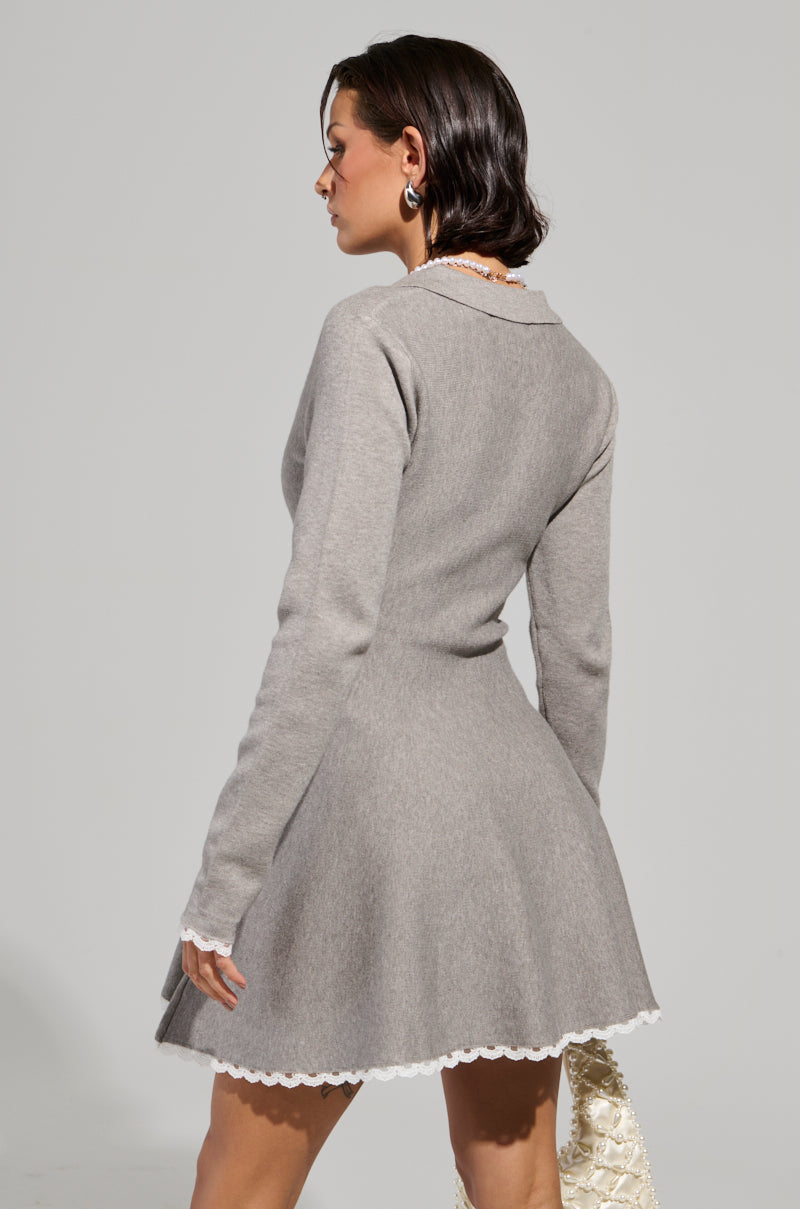 A person with short hair stands facing away, wearing a gray IN THE DETAILS KNIT MINI DRESS with lace trim and a flared skirt. They hold a beige handbag and wear hoop earrings, set against a neutral background.