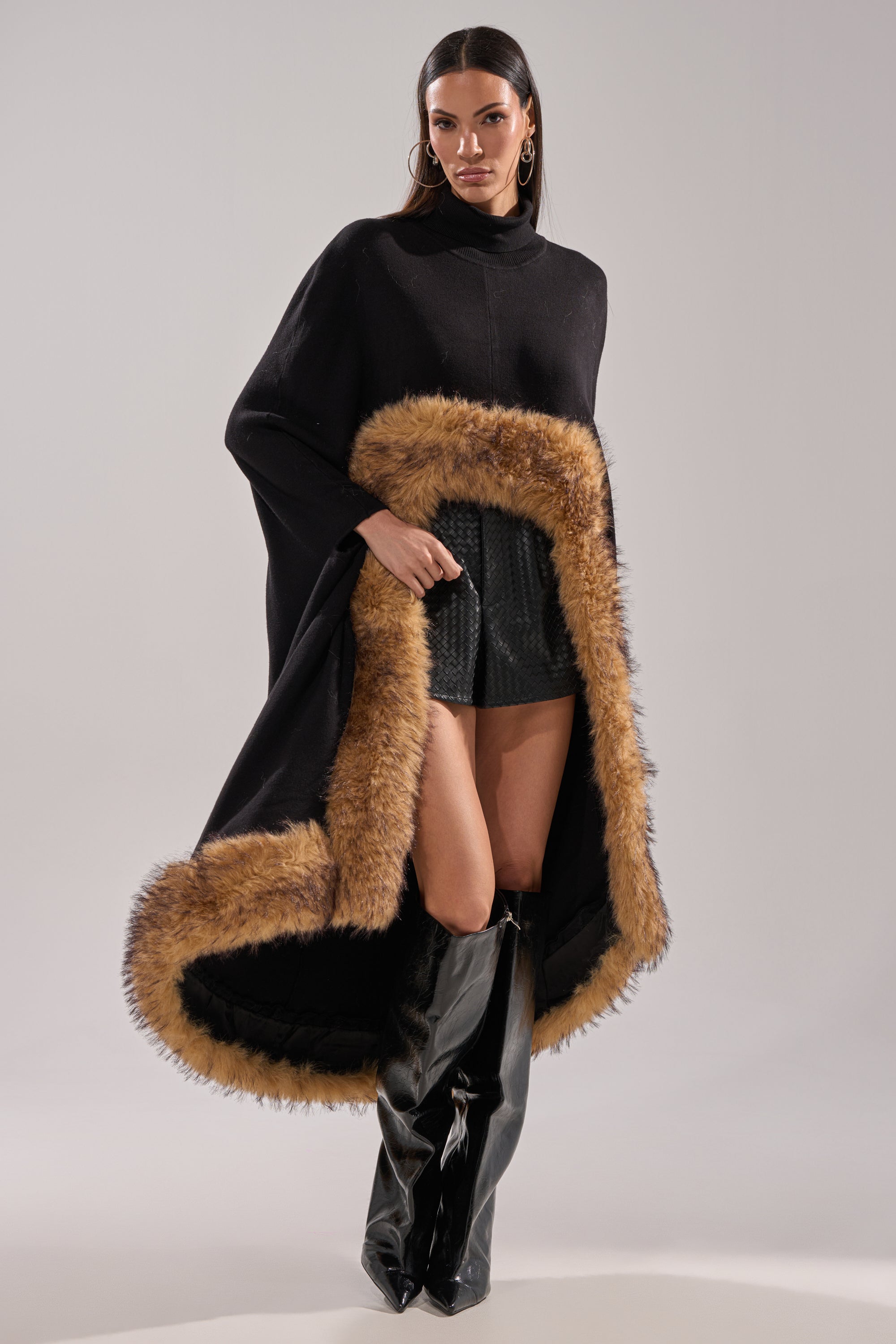 A woman models the RICH SWEATER WITH FUR TRIM, pairing it with a black skirt and tall boots against a light gray background for a stylish, modern winter layered look. 0