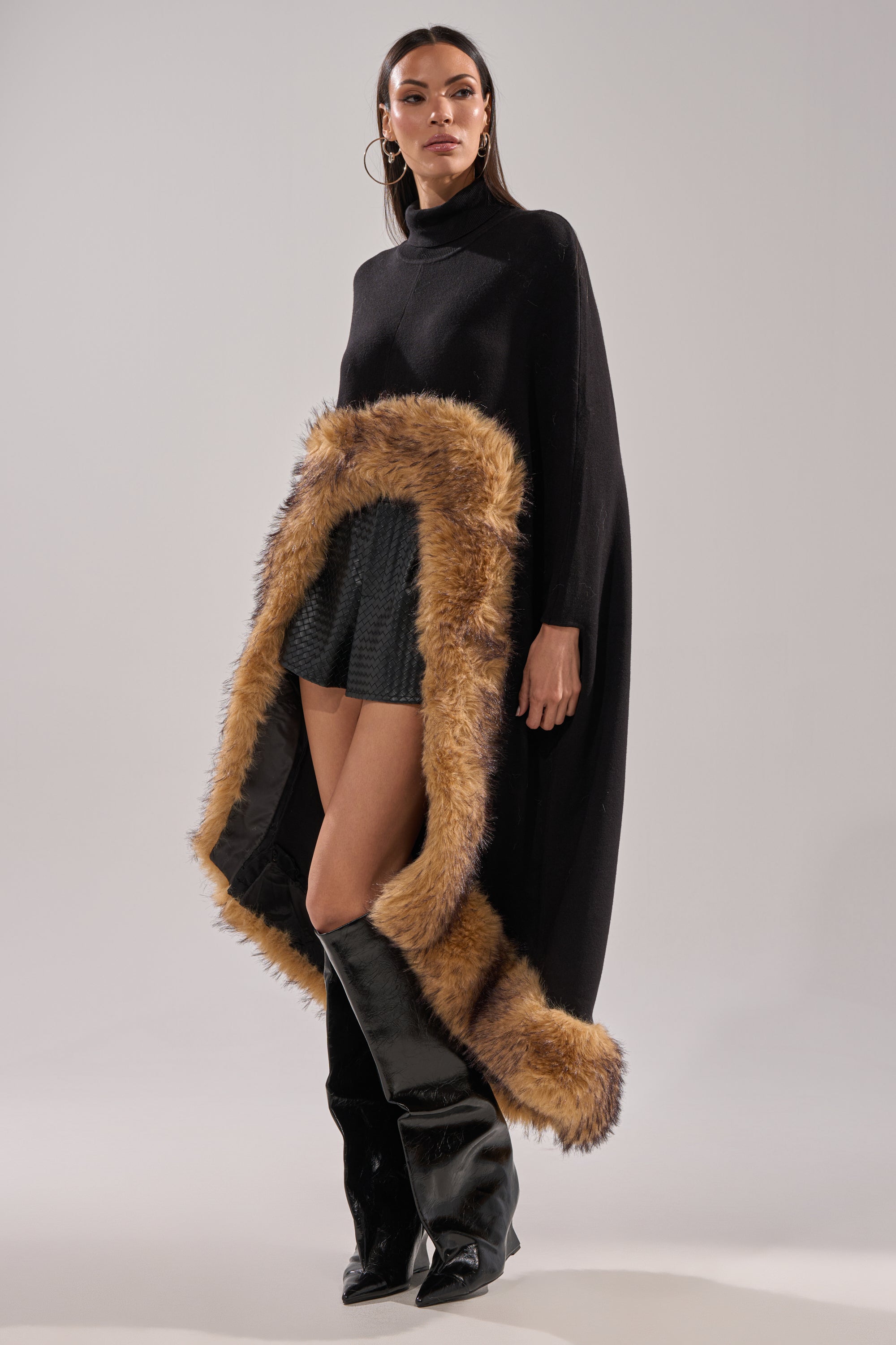 A woman models the RICH SWEATER WITH FUR TRIM as a black turtleneck poncho, paired with black textured shorts and shiny knee-high boots against a plain light background—a chic choice for winter layering. 1