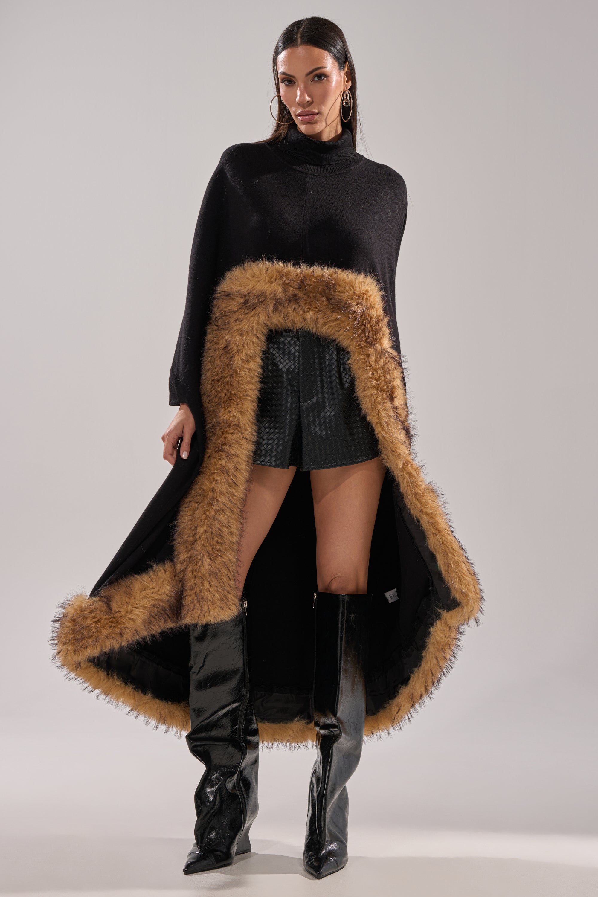 A woman confidently poses against a plain background wearing the RICH SWEATER WITH FUR TRIM as a winter layering piece, paired with black textured shorts and shiny black knee-high boots. 2