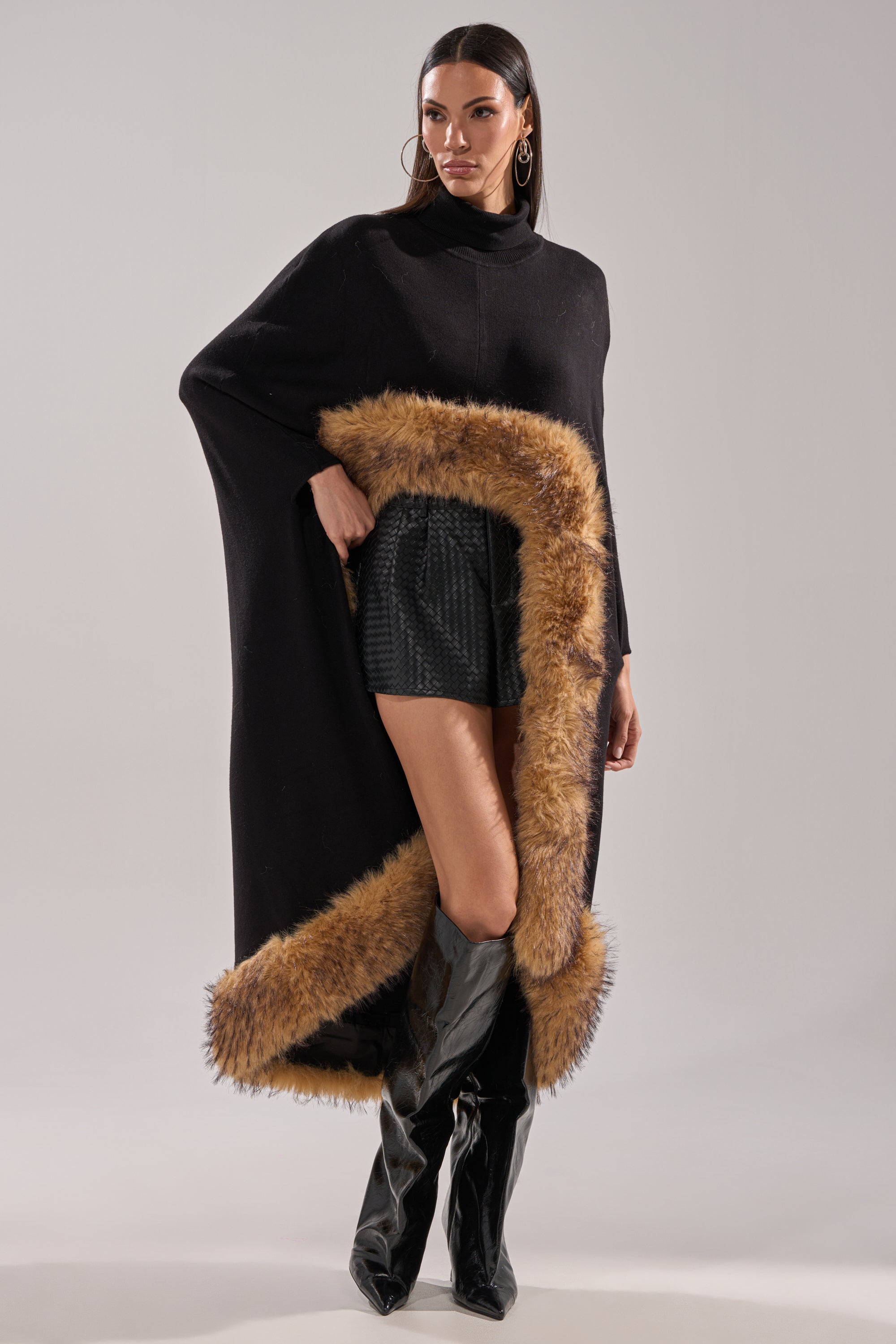 A woman showcases the RICH SWEATER WITH FUR TRIM, pairing it with a black textured mini skirt and knee-high black boots for a confident winter look against a plain background. 3