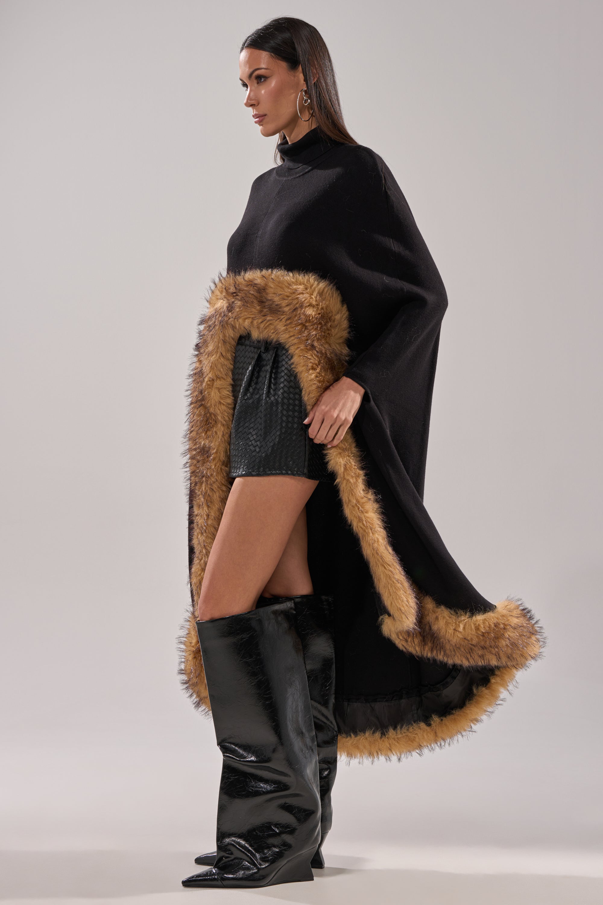 A woman models the RICH SWEATER WITH FUR TRIM, styled with black textured mini shorts and shiny black knee-high boots, set against a plain light background. 4