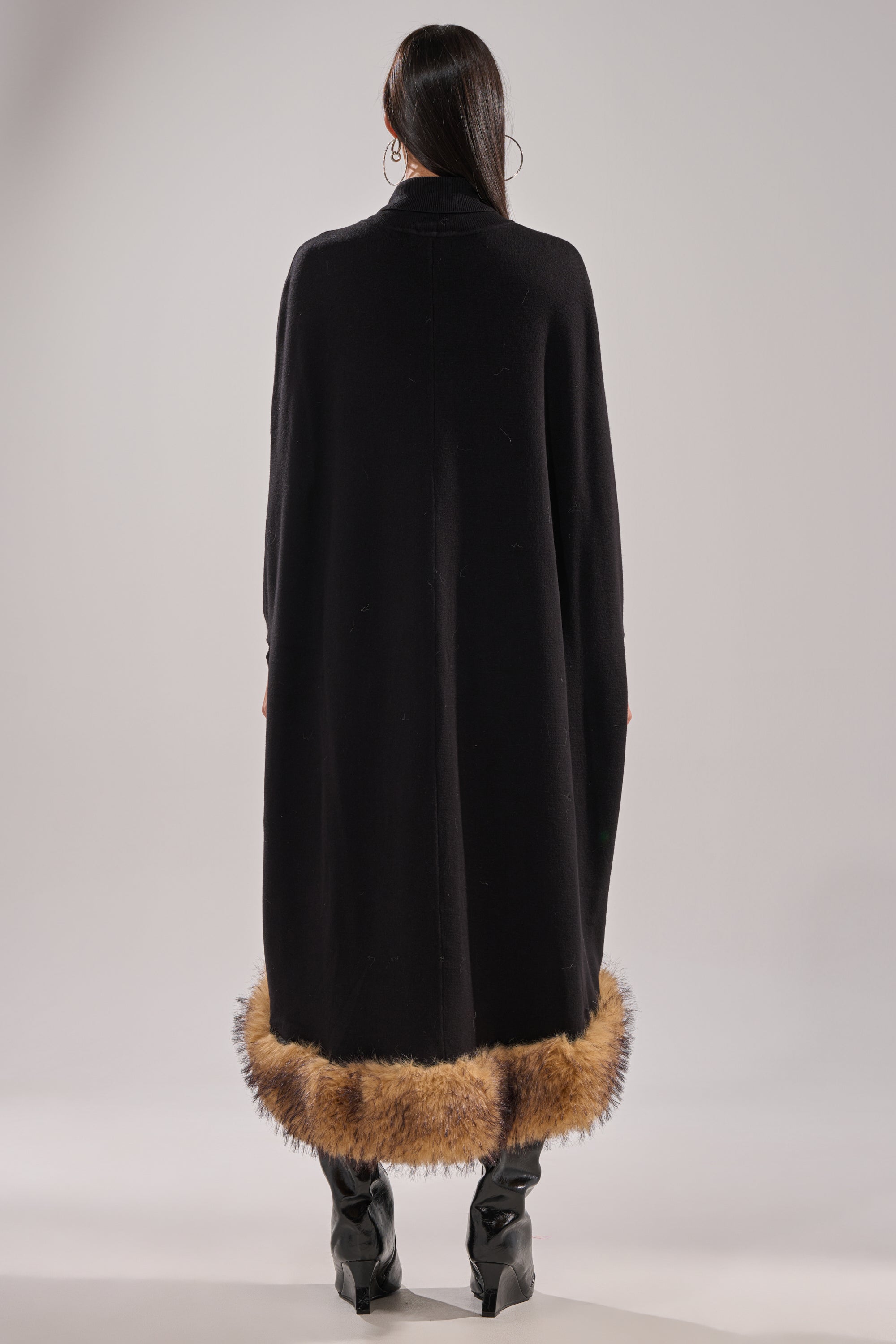 A person faces away, wearing the RICH SWEATER WITH FUR TRIM—a long black layering piece with brown fur at the hem—paired with black heeled boots and large hoop earrings, set against a light gray background. 6