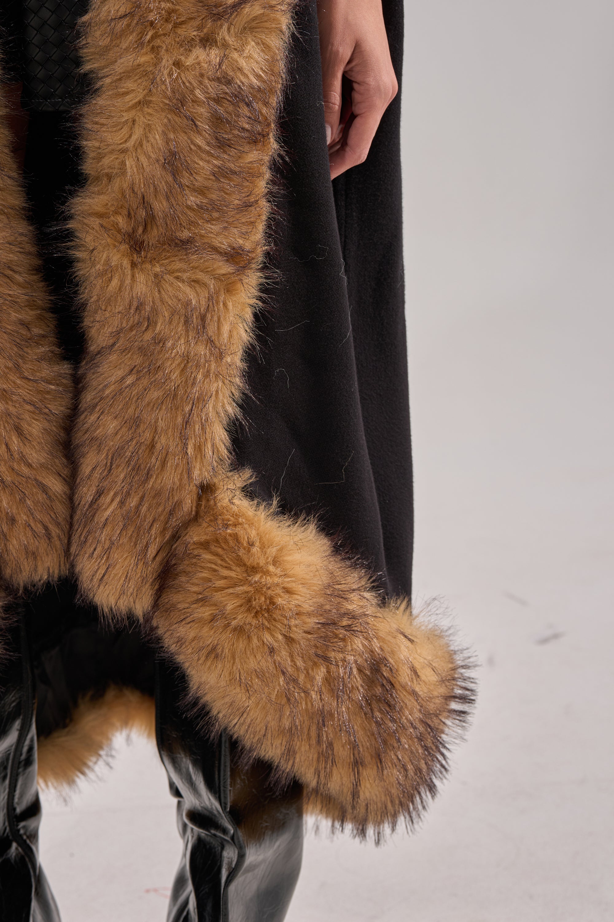 Wearing the RICH SWEATER WITH FUR TRIM, featuring thick light brown fur and paired with glossy black boots, only a hand, the lower part of the sweater, and boots are visible—creating an elegant winter look against a plain backdrop. 8