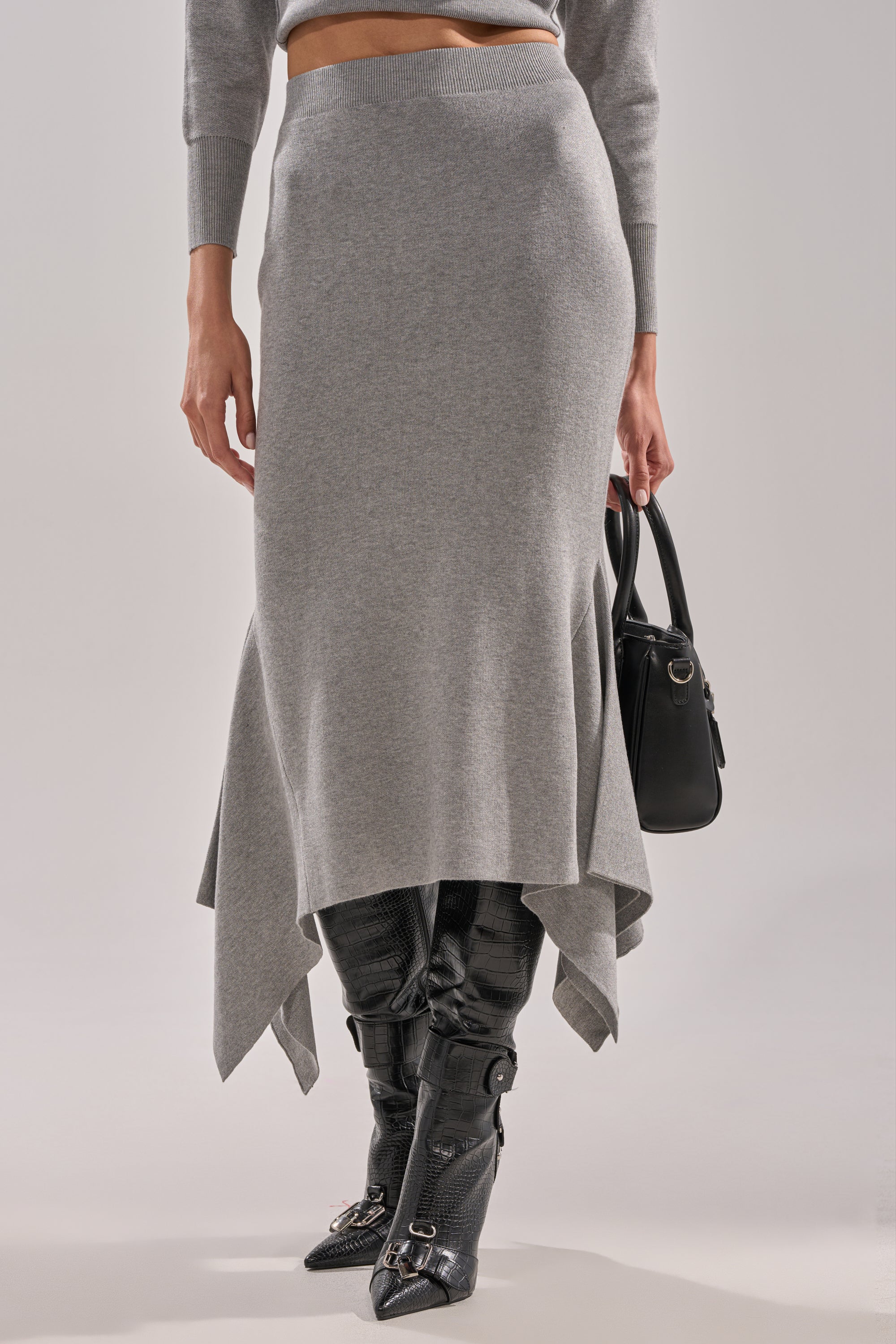 Wearing the LEXI MIDI SKIRT in gray with an asymmetrical hem and matching sweater, this person pairs their look with a black handbag and knee-high black heeled boots featuring buckle accents. 0