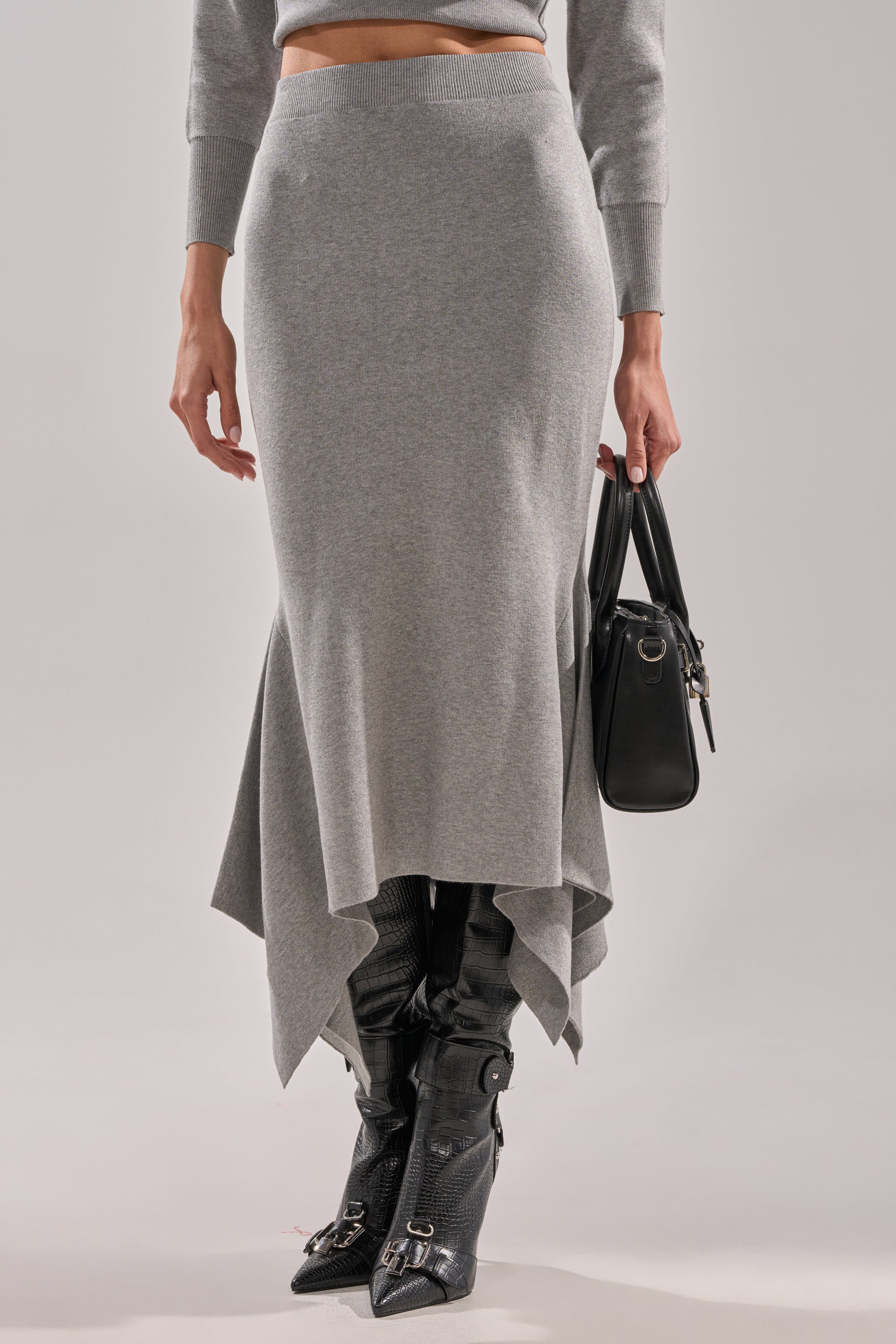 A person models the LEXI MIDI SKIRT with an asymmetrical hem, paired with a matching gray top, black textured knee-high buckle boots, and a black handbag against a neutral background. 1